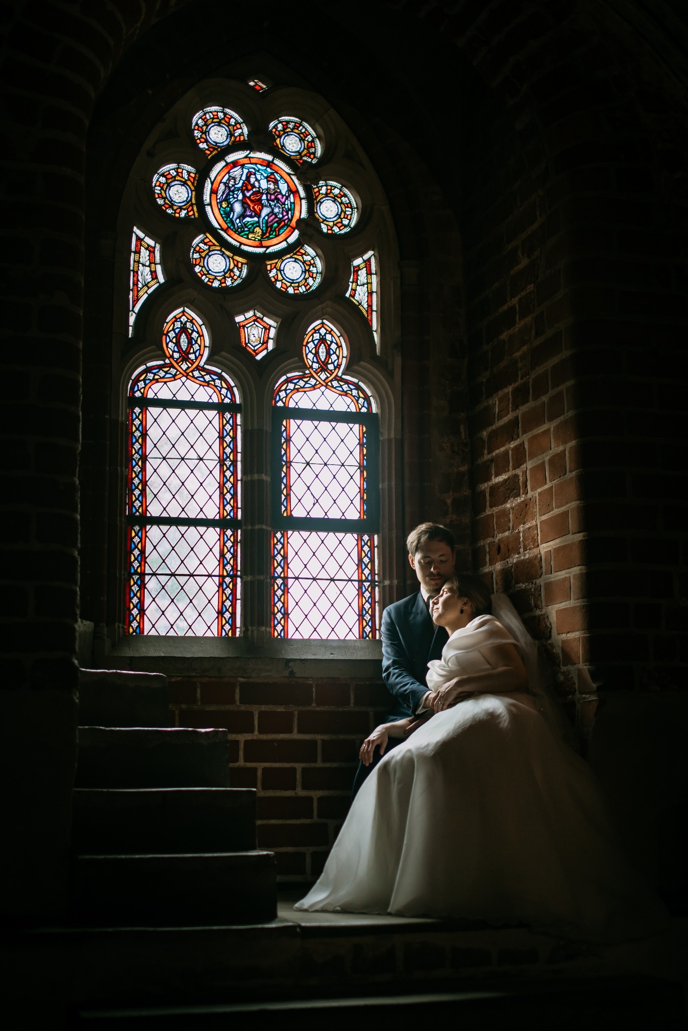 Para młoda w gotyckim wnętrzu, siedząca na schodach przy witrażowym oknie. Ciemne cegły i stonowane światło tworzą intymną atmosferę. Suknia panny młodej kontrastuje z tłem.