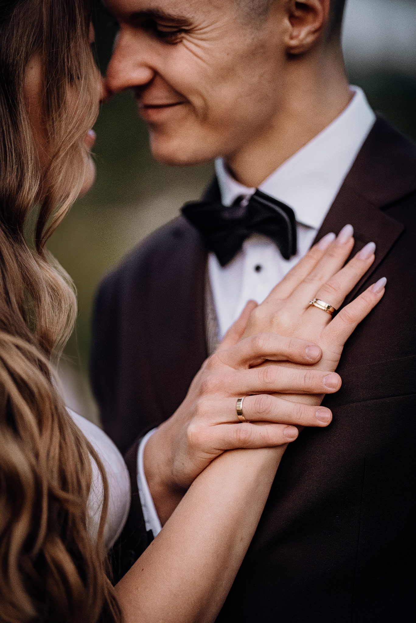 Zbliżenie dłoni pary młodej z obrączkami na tle garnituru i sukni. Delikatne światło podkreśla detale, tworząc intymną atmosferę.