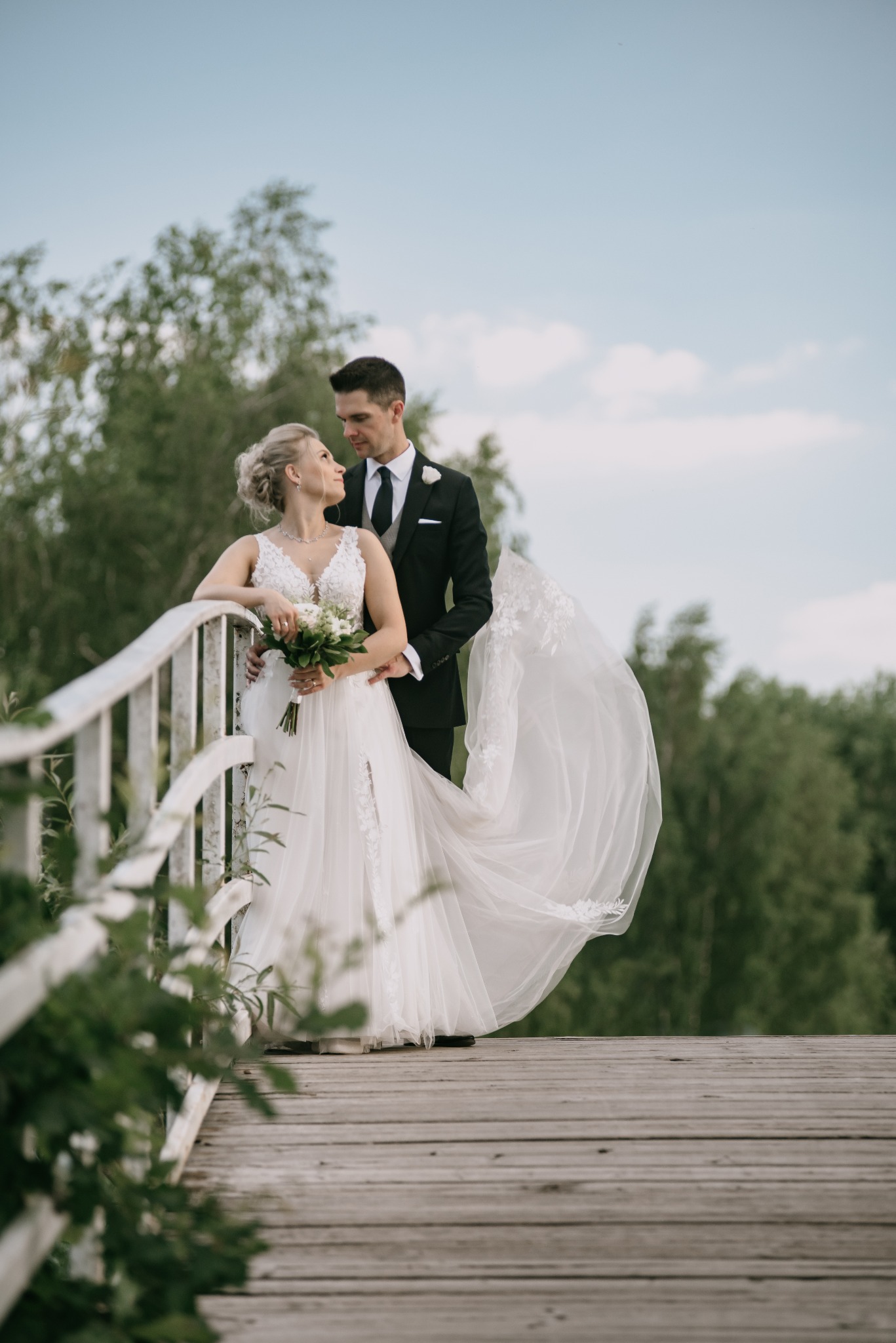 Para młoda na drewnianym moście, panna młoda w białej sukni z bukietem, pan młody w czarnym garniturze, delikatne oświetlenie, zielone tło drzew.