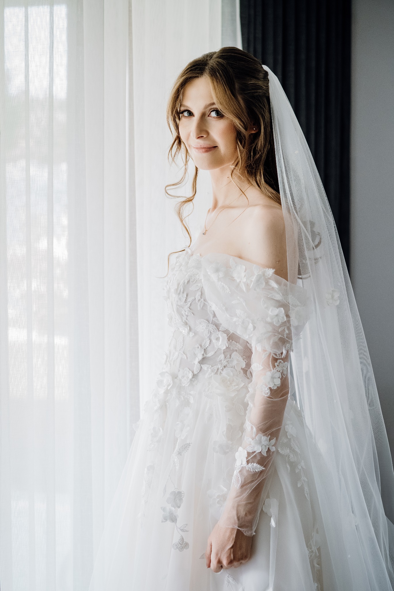 Panna młoda w białej sukni z kwiatowymi aplikacjami i długim welonem, pozująca przy oknie z delikatnymi firankami. Subtelny uśmiech, naturalny makijaż i rozpuszczone włosy.