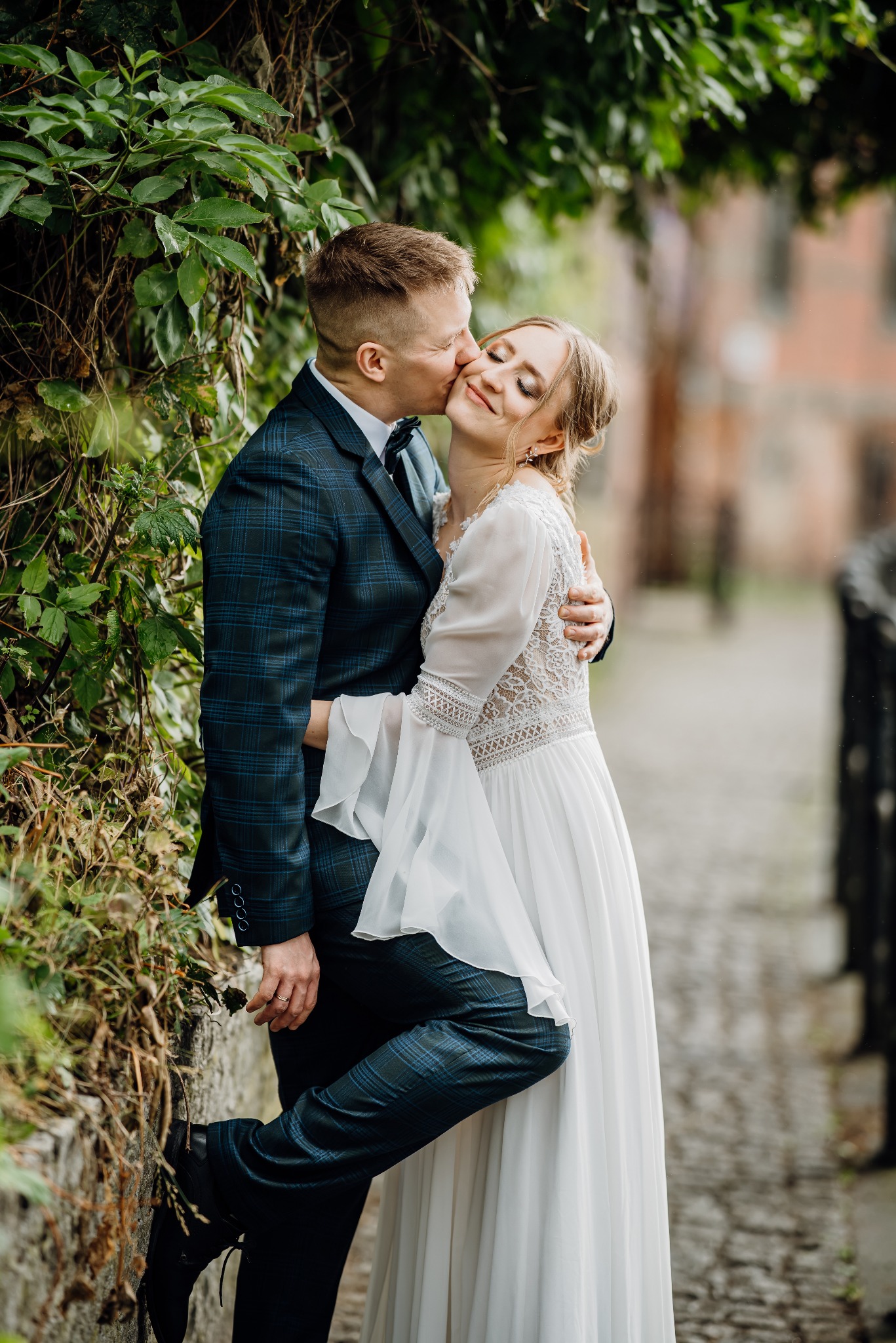 Panna młoda w zwiewnej sukni z koronką, z zamkniętymi oczami, całowana w policzek przez pana młodego w kraciastym garniturze na tle zieleni i muru.