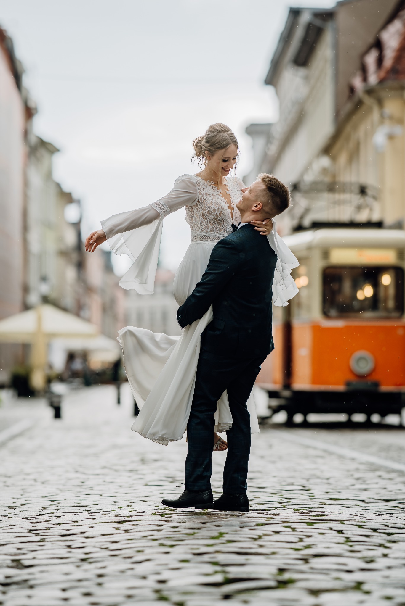 Uśmiechnięta panna młoda w koronkowej sukni, podnoszona przez pana młodego na tle brukowanej ulicy i pomarańczowego tramwaju w deszczowy dzień.