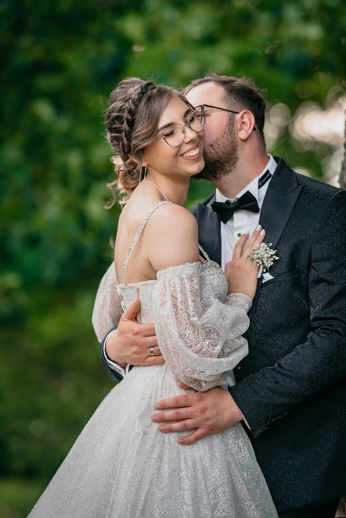 Uśmiechnięta panna młoda w okularach, obejmowana przez pana młodego w Bydgoszczy, podczas romantycznego pocałunku na tle zieleni.