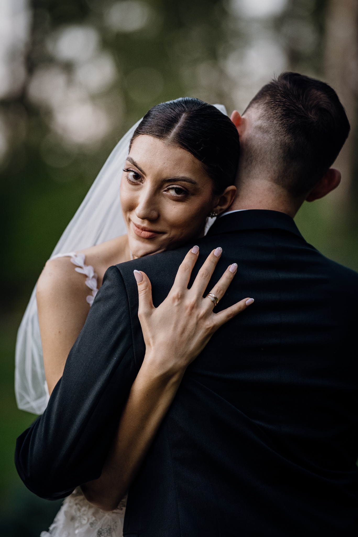 Panna młoda w objęciach pana młodego, z uśmiechem spogląda w obiektyw, prezentując pierścionek zaręczynowy na tle zieleni.