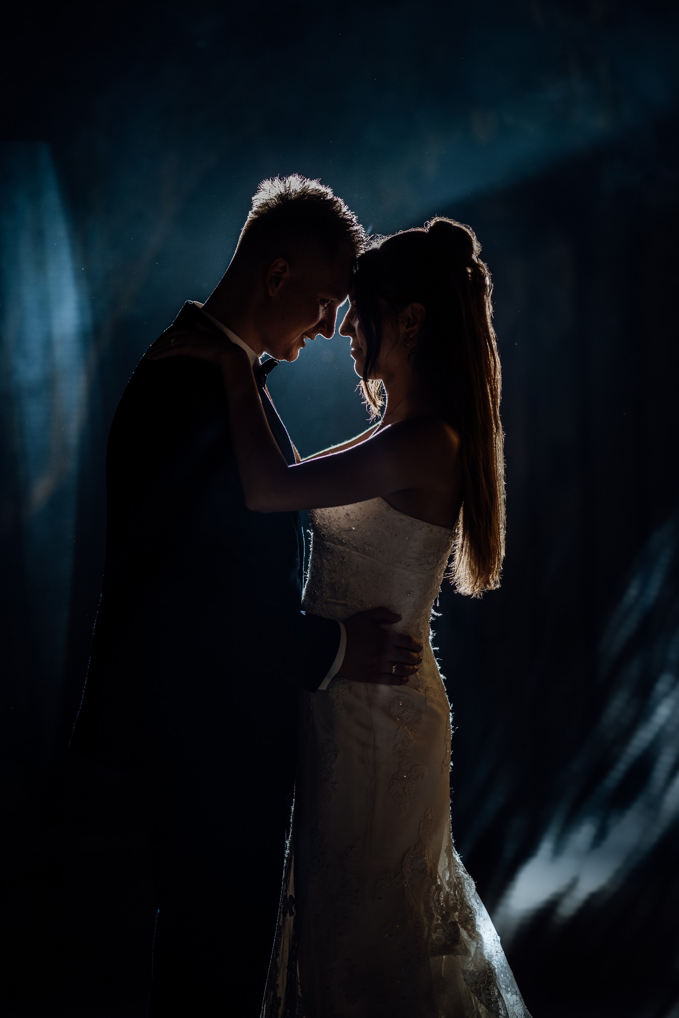 Silhouetted para młoda w objęciach, oświetlona od tyłu, tworząc romantyczny nastrój. Suknia panny młodej z delikatnymi zdobieniami. Ciemne tło z subtelnymi smugami światła.