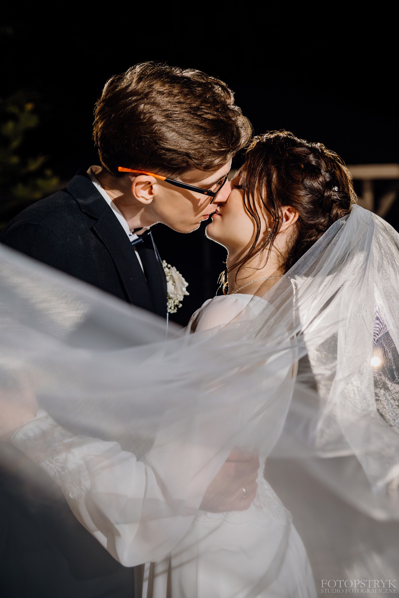 Młoda para w czułym pocałunku, on w okularach, ona w welonie, ciemne tło, ciepłe oświetlenie, delikatny ruch welonu, intymna atmosfera, fotografia ślubna.