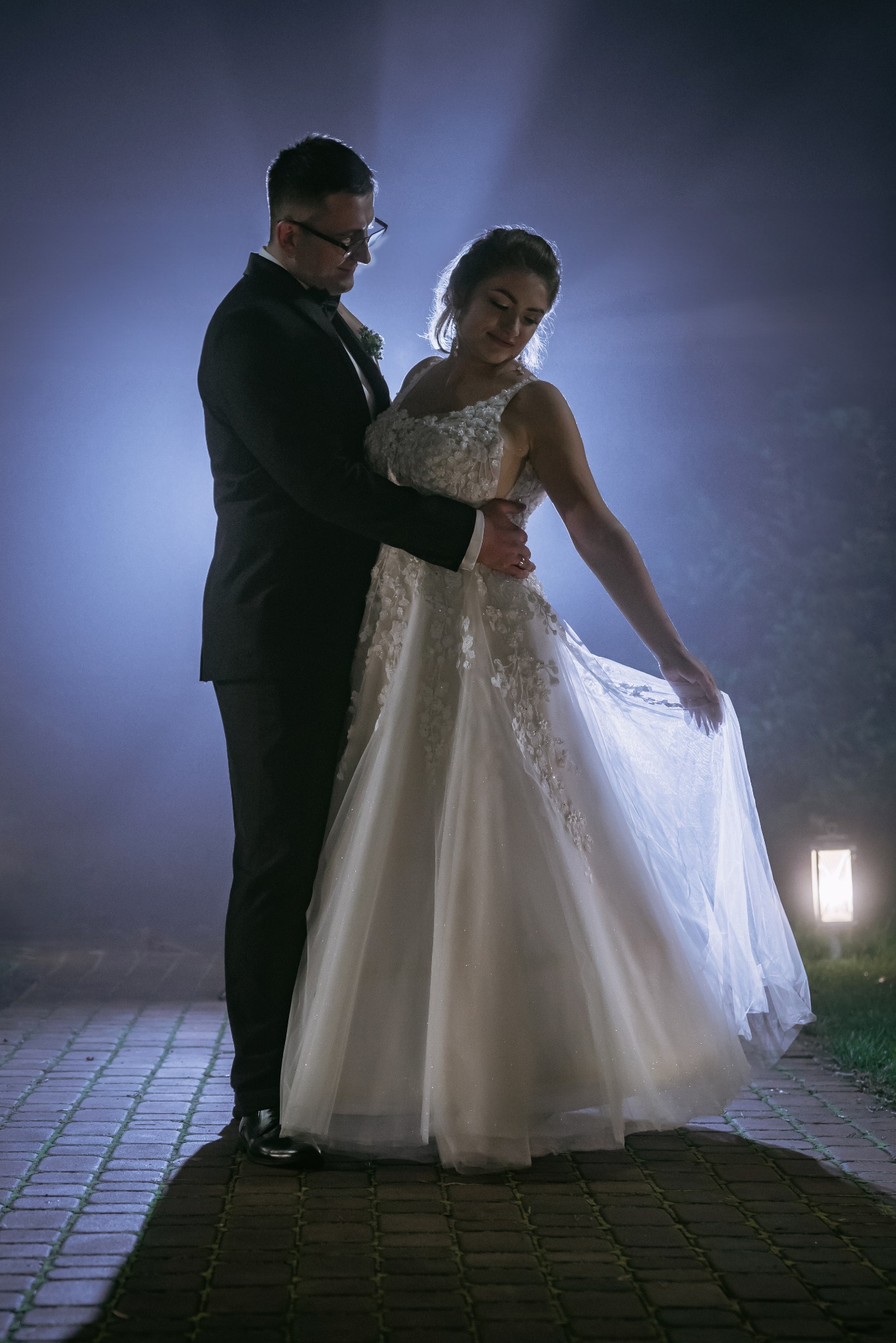 Para młoda w objęciach na tle nocnego, lekko zamglonego ogrodu z subtelnym oświetleniem. Panna młoda w białej sukni z koronkowymi aplikacjami, pan młody w czarnym smokingu.