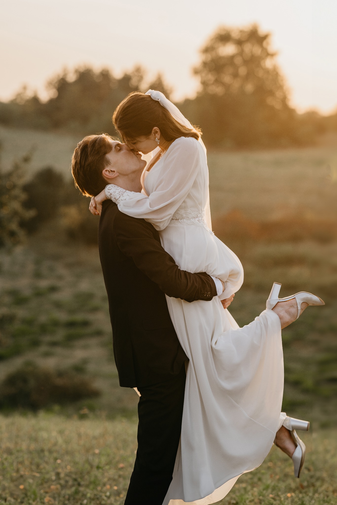 Para młoda w objęciach na tle zachodzącego słońca. Panna młoda w zwiewnej sukni i welonie, trzymana przez pana młodego. Romantyczna atmosfera, ciepłe światło.