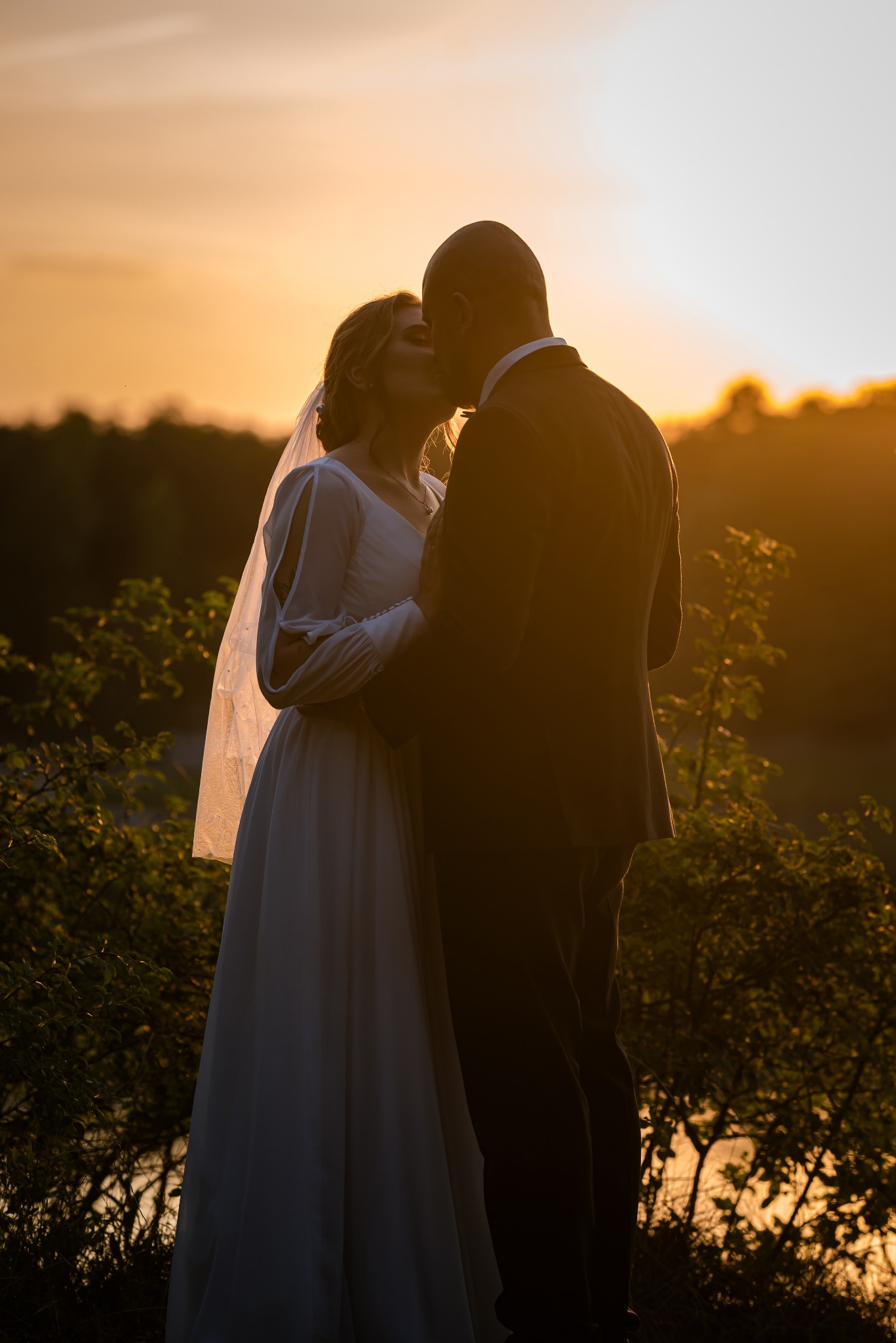 Silhouetted para młoda całuje się na tle zachodzącego słońca. Romantyczna sceneria z naturalnym światłem i delikatną sylwetką drzew w tle.