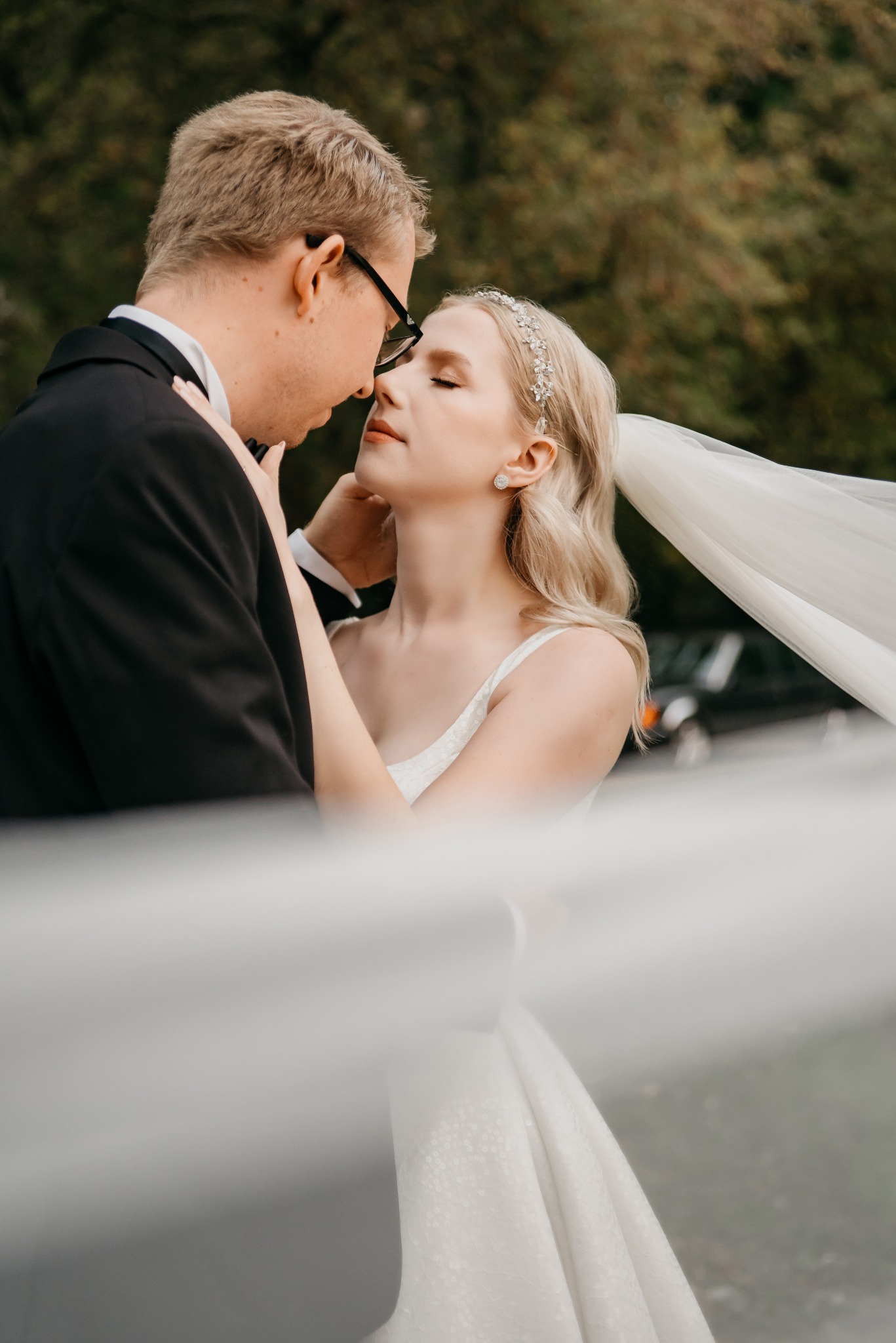 Delikatne zbliżenie na parę młodą w objęciach, welon w tle, subtelne światło podkreśla emocje, drzewa w tle, on w czarnym smokingu, ona w białej sukni.