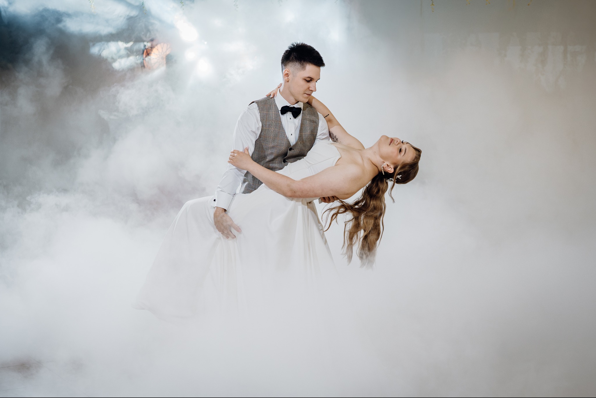 Para młoda w tańcu, on w kamizelce w kratę, ona w białej sukni, otoczeni mgłą, subtelne oświetlenie podkreśla romantyczny nastrój.
