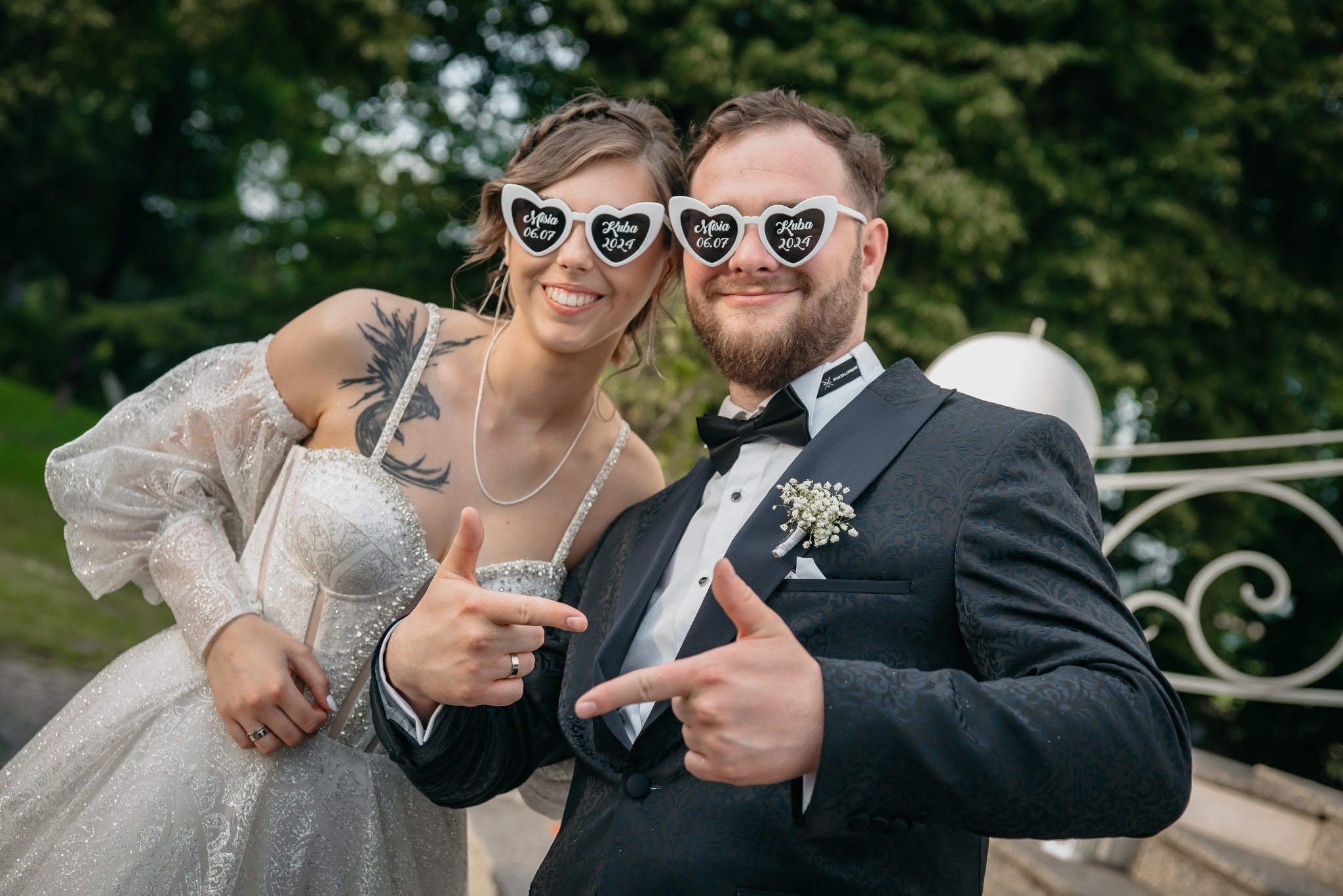 Uśmiechnięta para młoda w okularach w kształcie serc z datą ślubu, pozuje na tle zieleni. Pan młody w eleganckim smokingu, panna młoda w błyszczącej sukni.