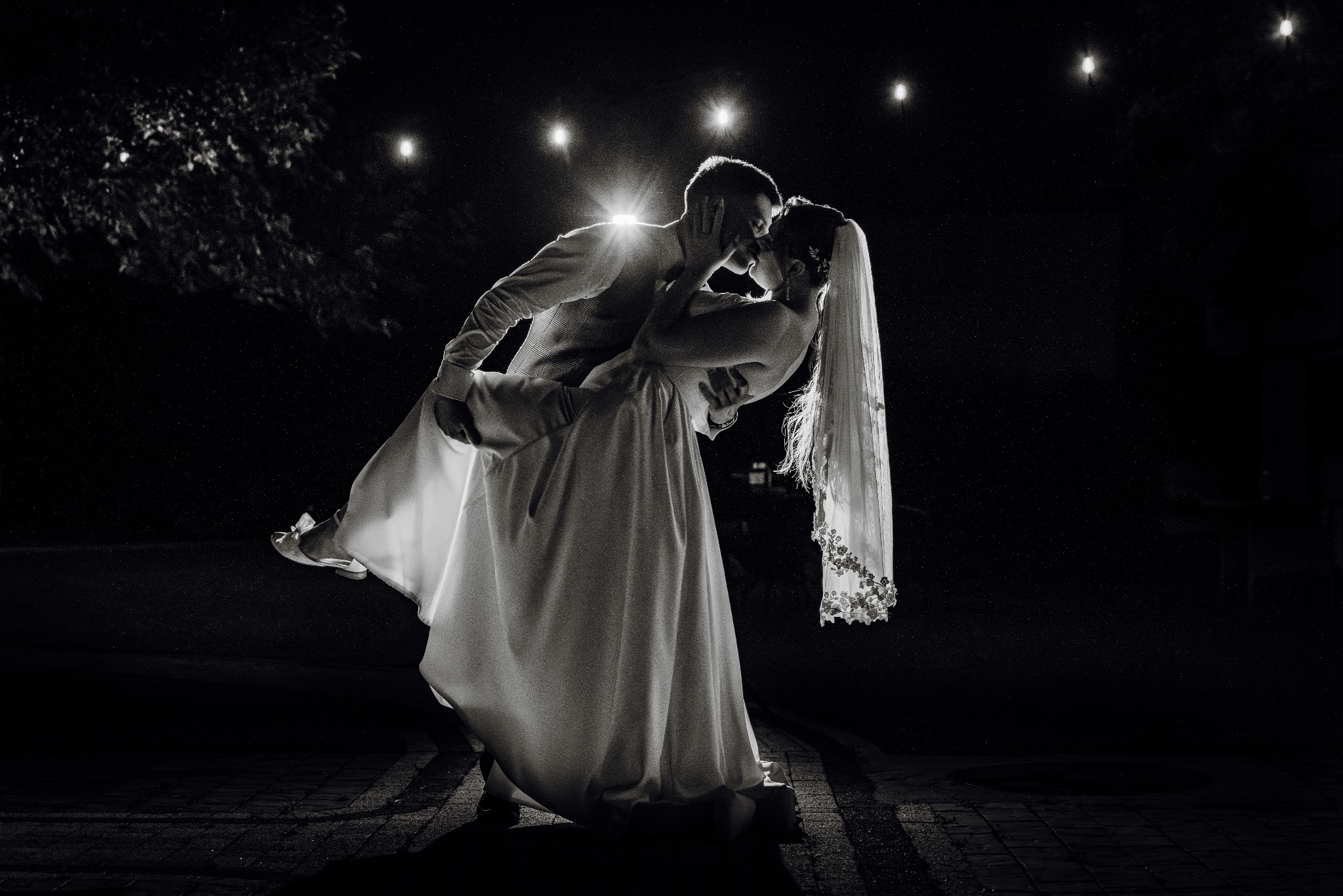 Czarno-białe zdjęcie pary młodej w pocałunku, panna młoda w sukni i welonie, pan młody trzyma pannę młodą w ramionach, oświetlenie z tyłu tworzy efektowny blask.