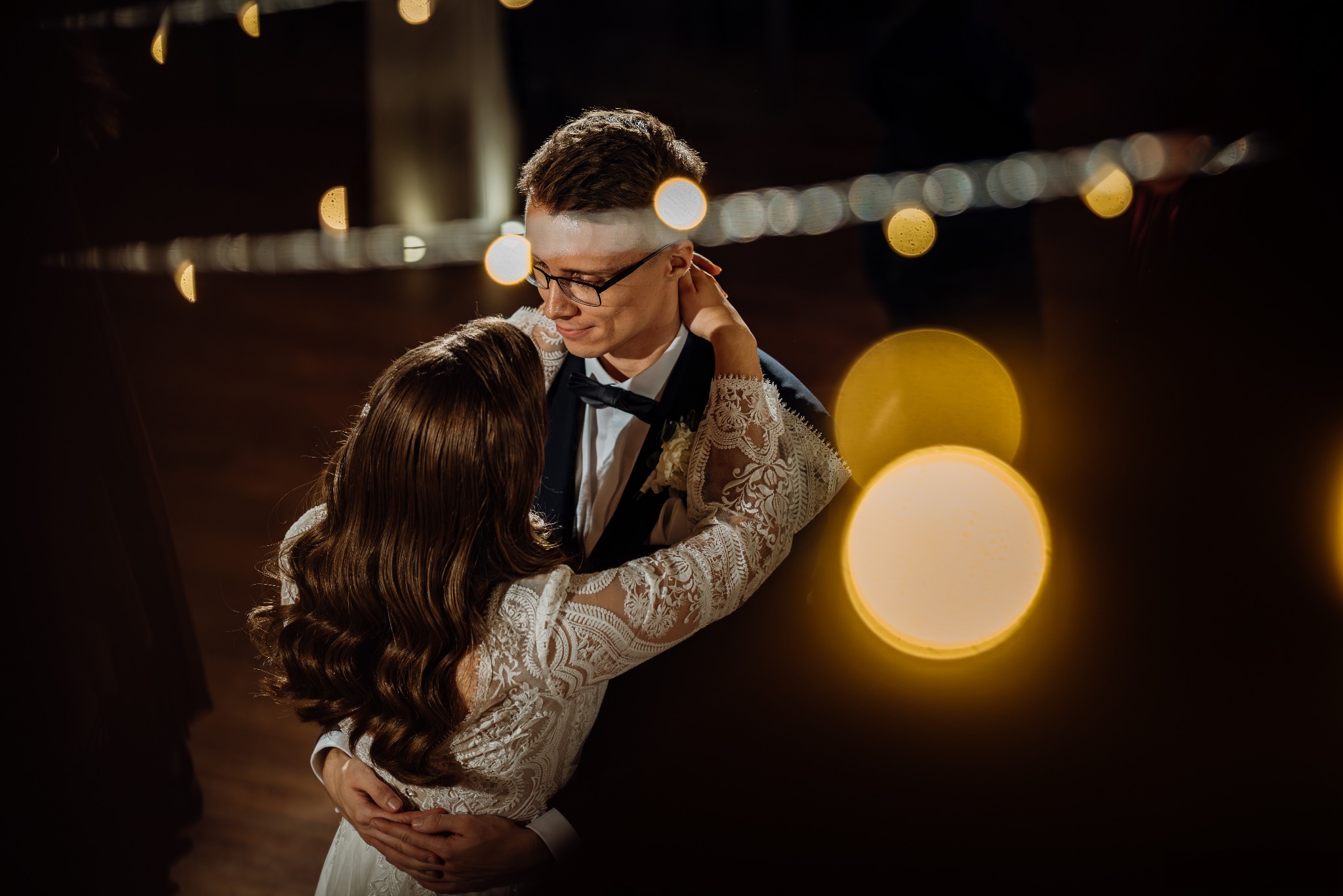 Para młoda w objęciach, on w ciemnym garniturze i muszce, ona w koronkowej sukni, ciepłe światło bokeh w tle, intymna chwila.