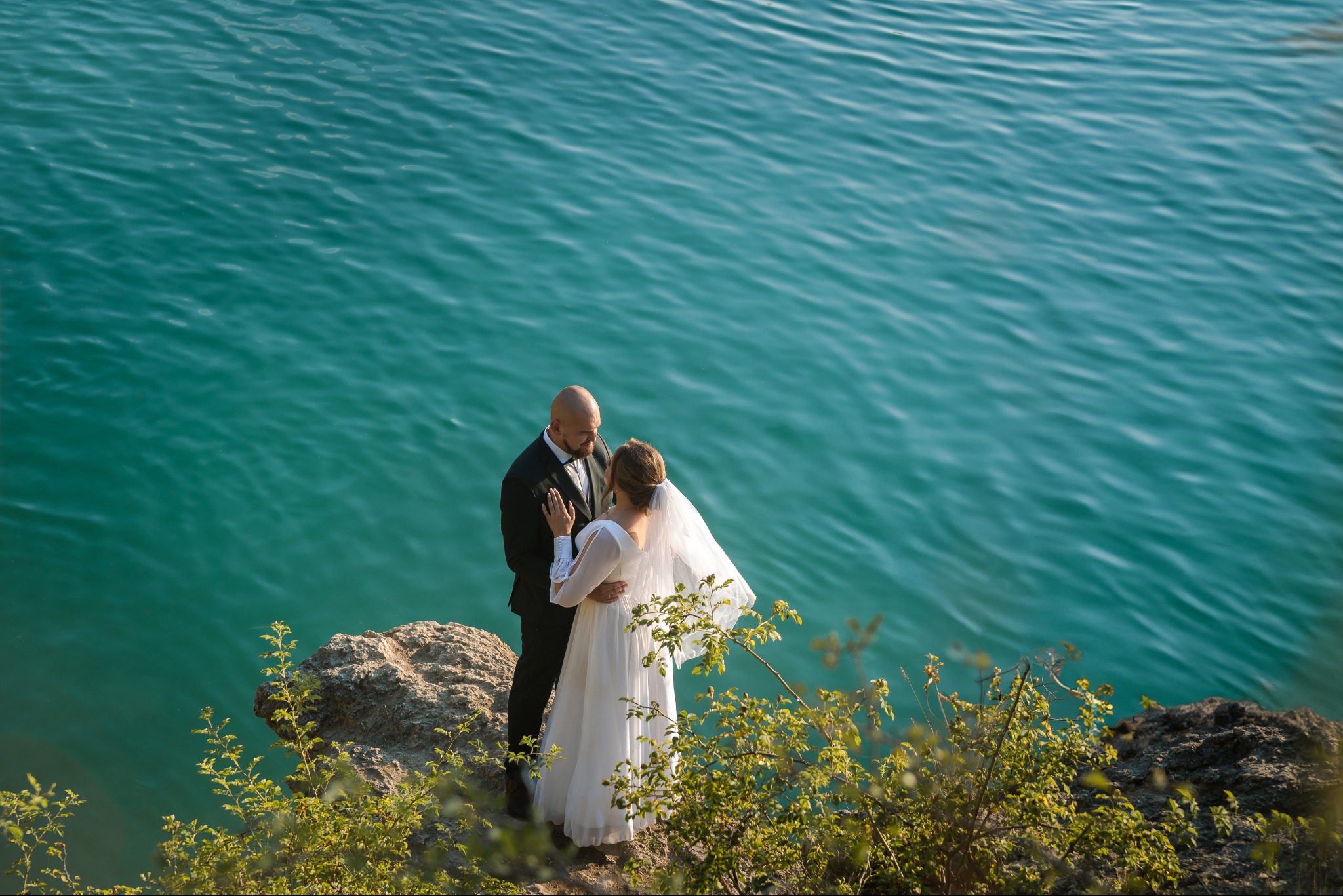 Para młoda w objęciach na tle turkusowej wody. Pan młody w czarnym smokingu, panna młoda w białej sukni z welonem. Romantyczna sesja plenerowa na skalistym brzegu.