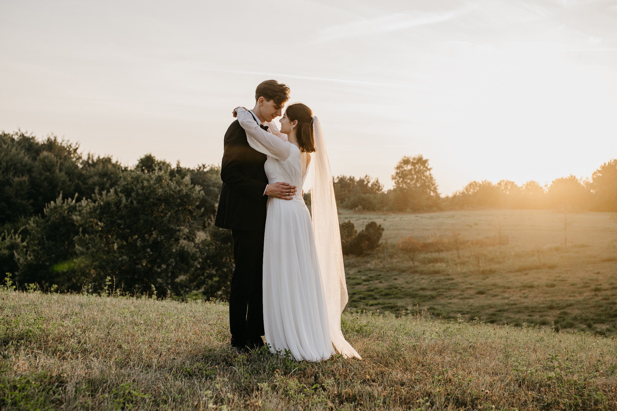 Para młoda w objęciach na tle zachodzącego słońca. Panna młoda w prostej sukni i welonie, pan młody w czarnym smokingu. Romantyczna sesja w plenerze.