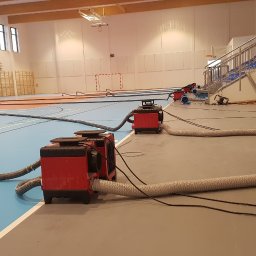 PHU OPTIMAA ŁUKASZ POLEROWICZ - Osuszanie podposadzkowe hali sportowej: czerwone osuszacze z szarymi wężami na parkiecie i szarej posadzce, widoczne trybuny i drabinki gimnastyczne.