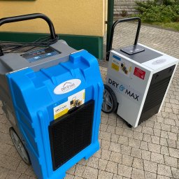 PHU OPTIMAA ŁUKASZ POLEROWICZ - Dwa profesjonalne urządzenia do osuszania budynków marki Dry & Max, stojące na kostce brukowej przed budynkiem. Jeden w kolorze niebieskim, drugi biały.