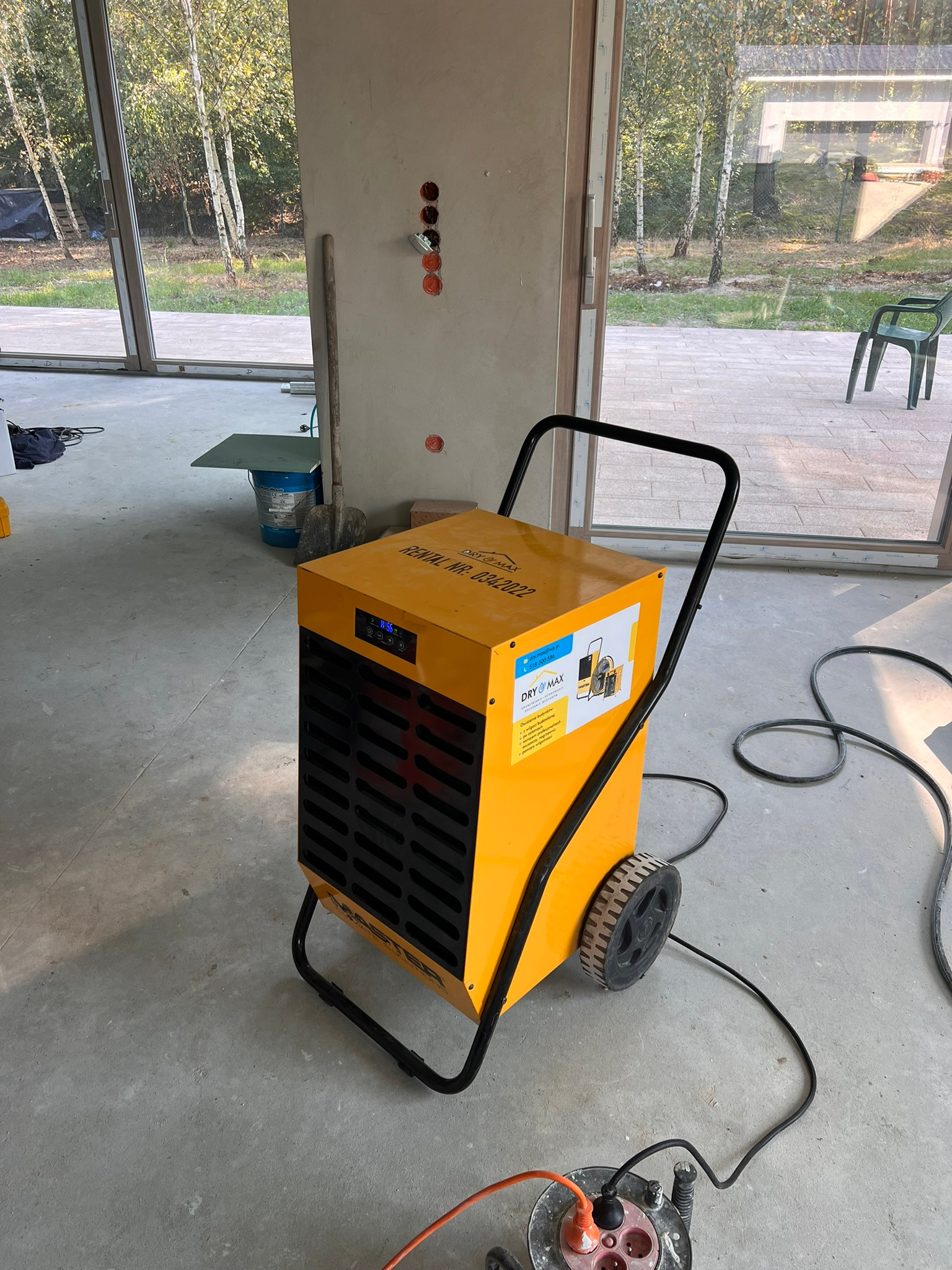 Żółty osuszacz budowlany DryMax na kółkach, z widocznymi przewodami, stoi w surowym wnętrzu z dużymi oknami, sugerującym etap prac wykończeniowych.