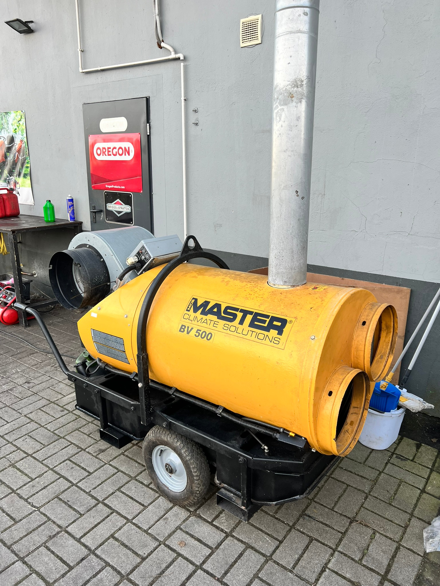 Żółta nagrzewnica Master BV 500 na kółkach, z kominem, stoi na kostce brukowej przed szarą ścianą z drzwiami i reklamą Oregon. Sprzęt do osuszania budynków.