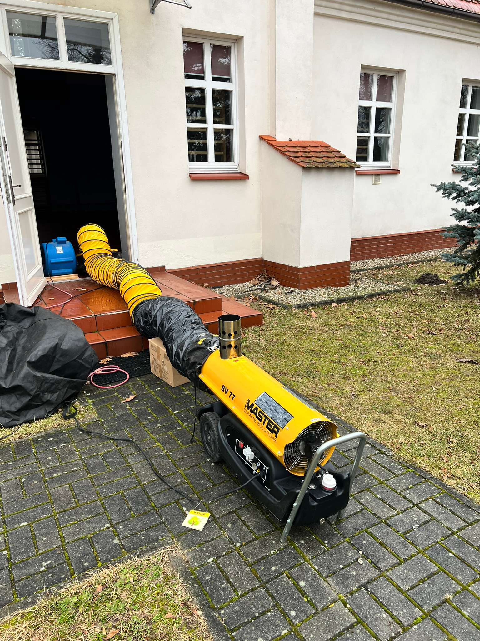 Żółty nagrzewnica MASTER BV 77 z podłączonym przewodem wentylacyjnym skierowanym do wnętrza budynku. Urządzenie stoi na kostce brukowej przed wejściem.