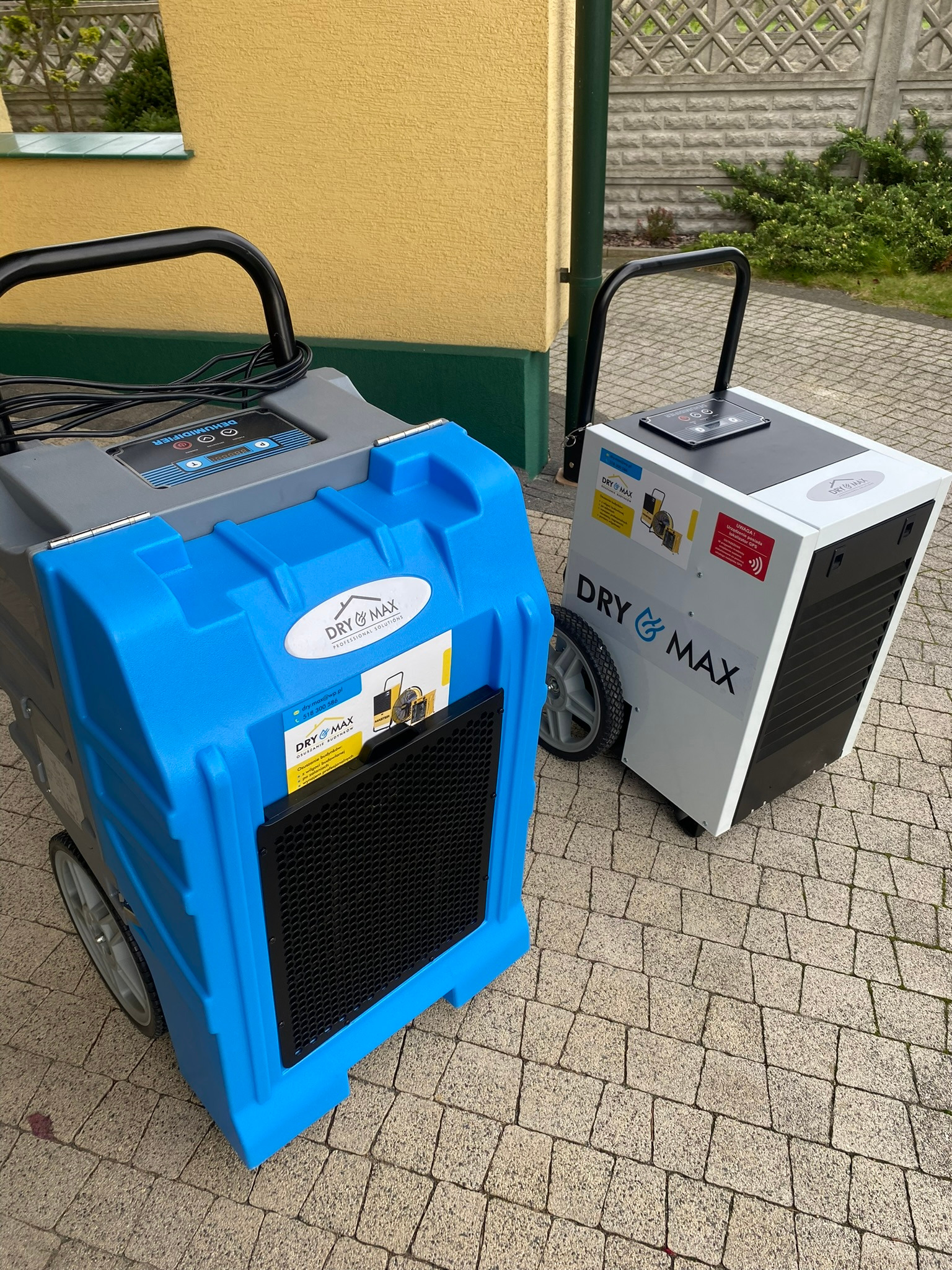 Dwa profesjonalne urządzenia do osuszania budynków marki Dry & Max, stojące na kostce brukowej przed budynkiem. Jeden w kolorze niebieskim, drugi biały.