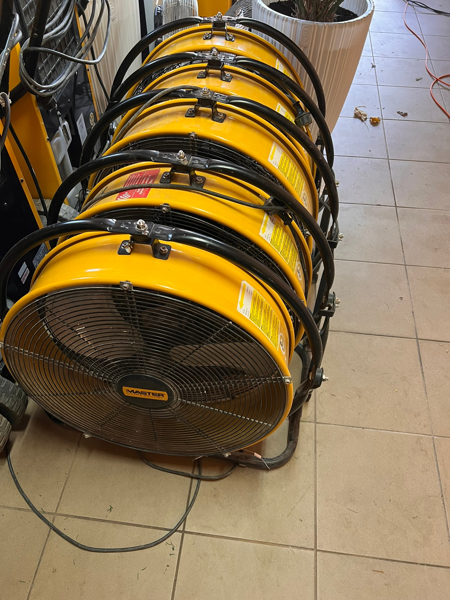 Żółte, przemysłowe wentylatory Master ustawione jeden na drugim, z widocznymi kablami i fragmentem donicy z rośliną w tle, na podłodze z płytek.