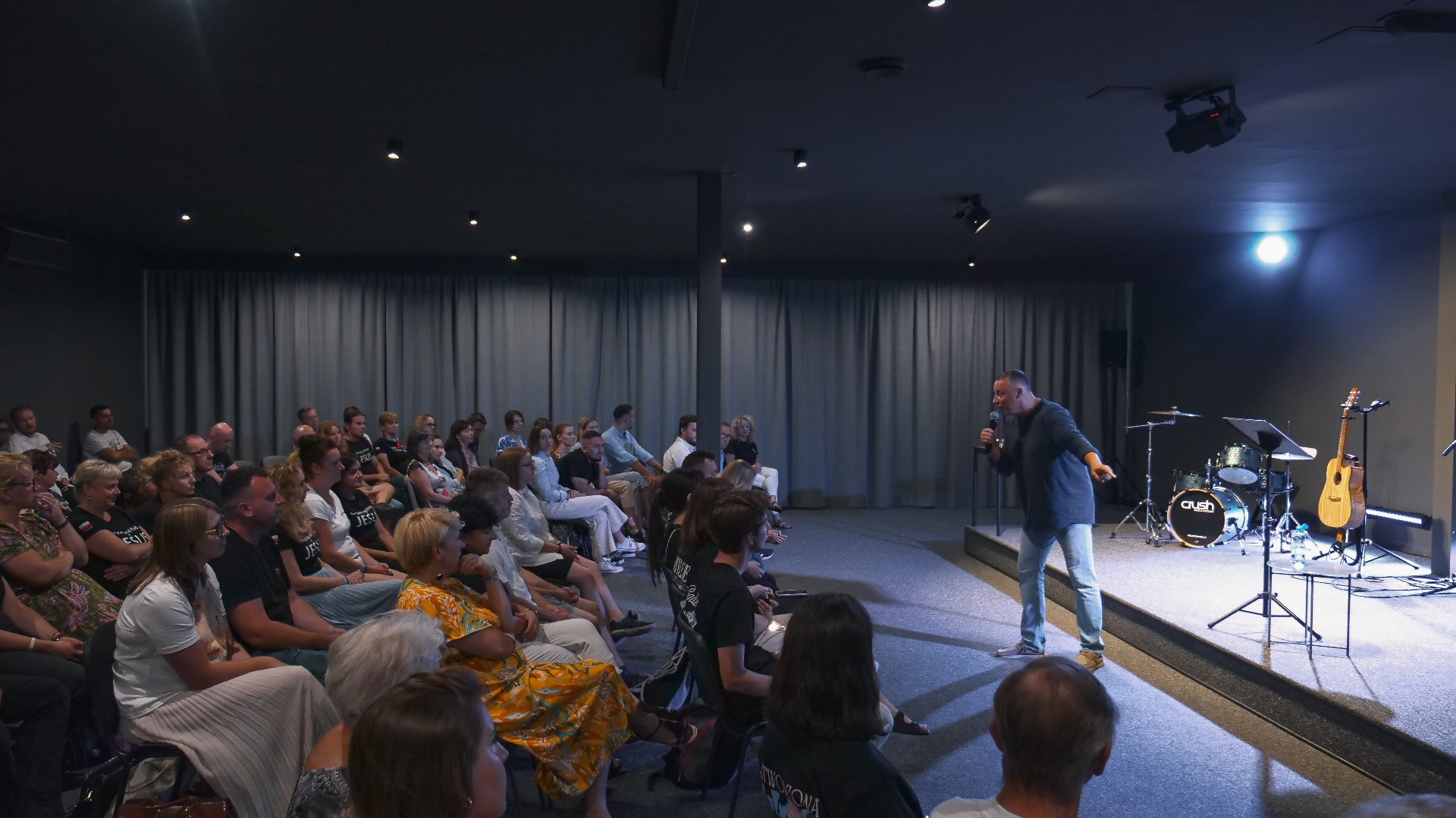 Prelegent z mikrofonem na scenie, publiczność siedzi na widowni. W tle instrumenty muzyczne i oświetlenie sceniczne. Ujęcie z perspektywy widza.