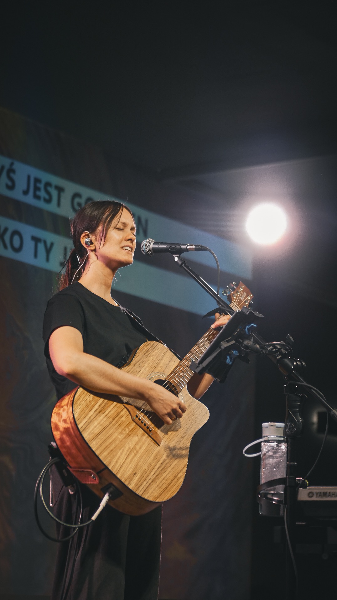 Kobieta z mikrofonem śpiewa i gra na gitarze akustycznej na scenie, oświetlona reflektorem. Widoczny keyboard Yamaha i fragment tekstu na banerze w tle.