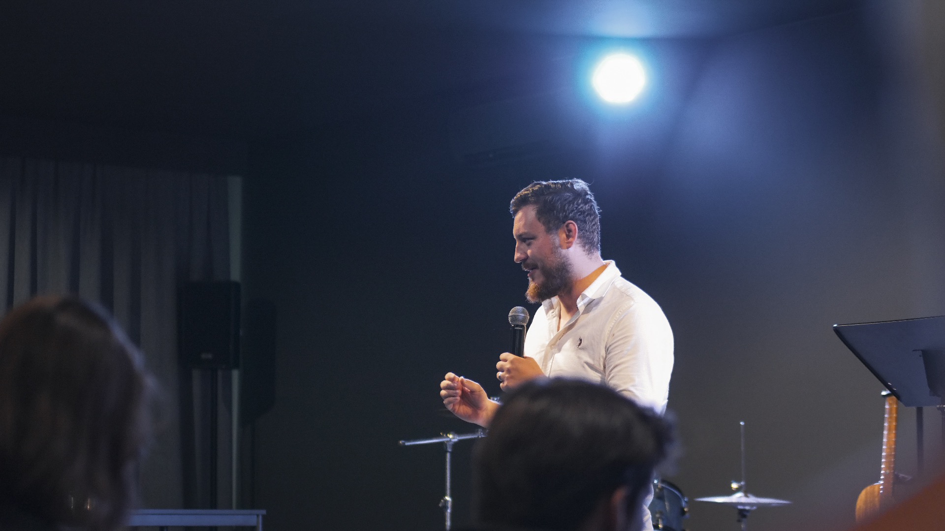 Mężczyzna z brodą w białej koszuli przemawia do mikrofonu na scenie, oświetlony reflektorem. W tle widoczni słuchacze i instrumenty muzyczne.
