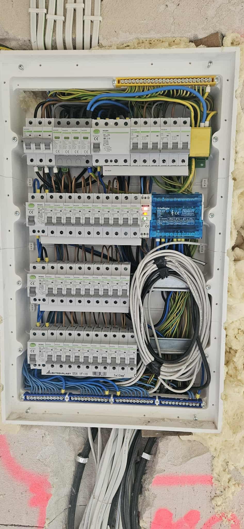 Rozdzielnica elektryczna w białej obudowie, widoczne zabezpieczenia, przewody w różnych kolorach, listwa zaciskowa. Ściana z pianką montażową w tle.