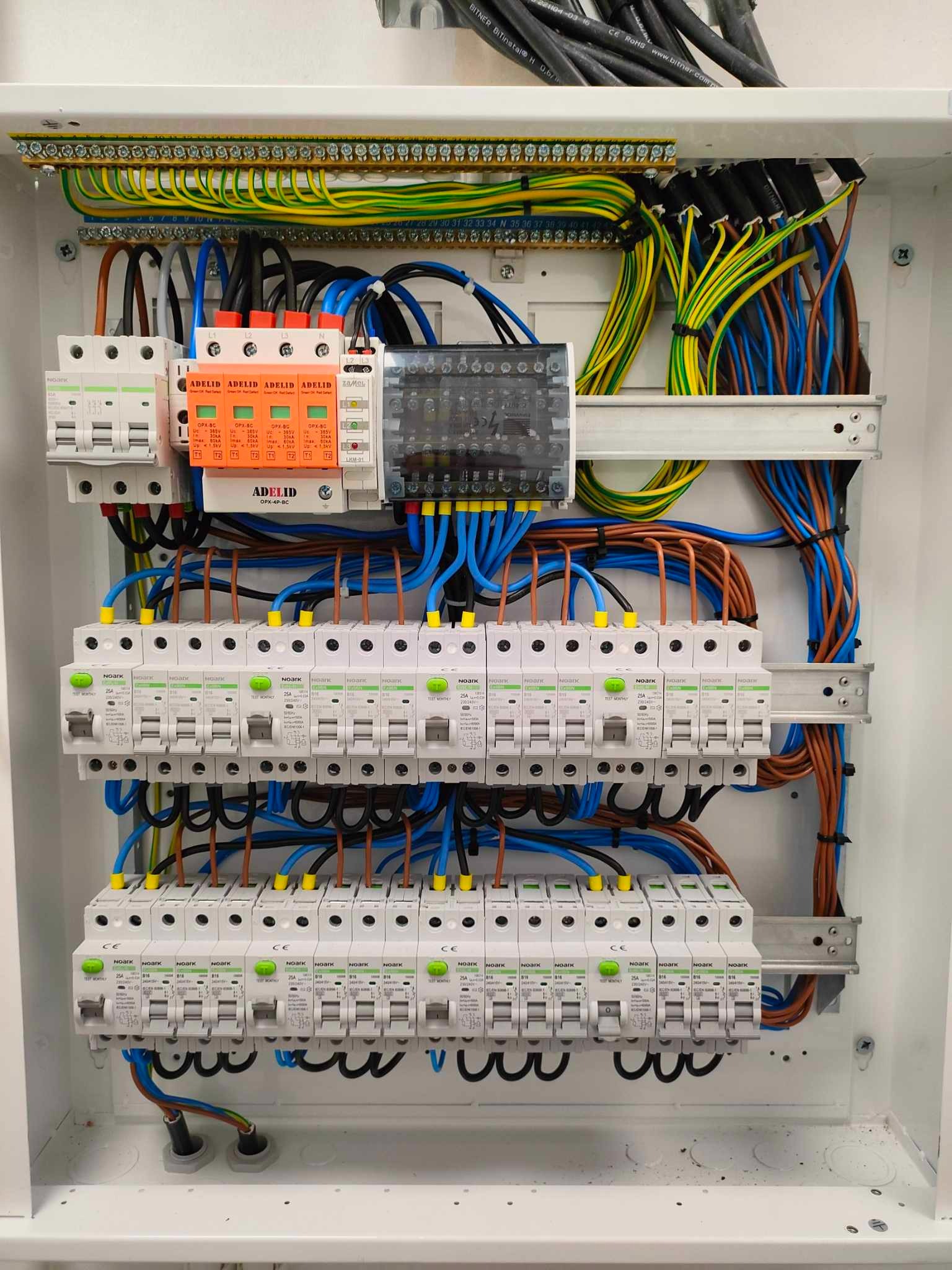 Rozdzielnia elektryczna z zainstalowanymi bezpiecznikami, przewodami w różnych kolorach i zabezpieczeniami przeciwprzepięciowymi marki Adelid. Widok z góry na otwartą skrzynkę.