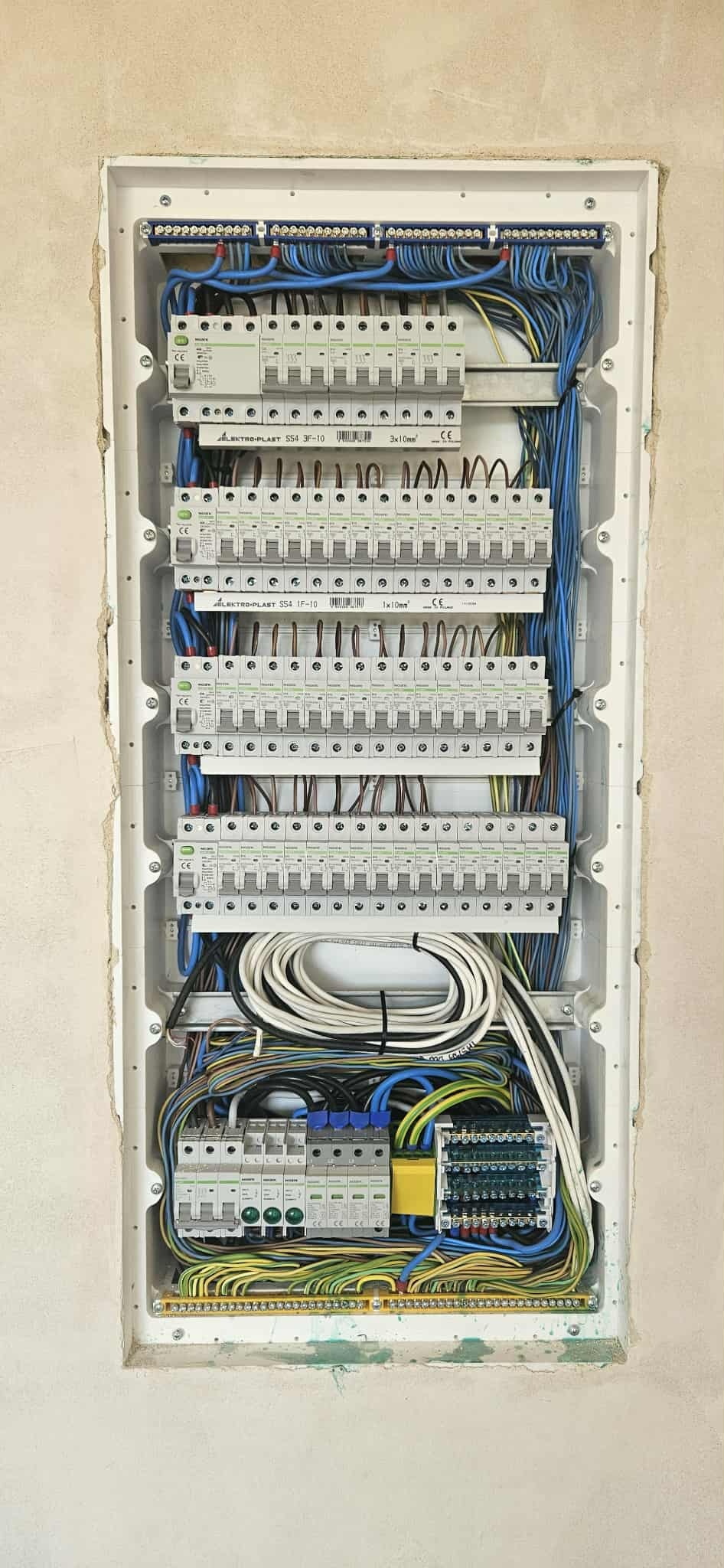 Rozdzielnica elektryczna wnękowa z licznymi bezpiecznikami, przewodami w kolorach: niebieskim, białym, żółtym i zielonym. Widoczne oznaczenia producenta Elektro-Plast.