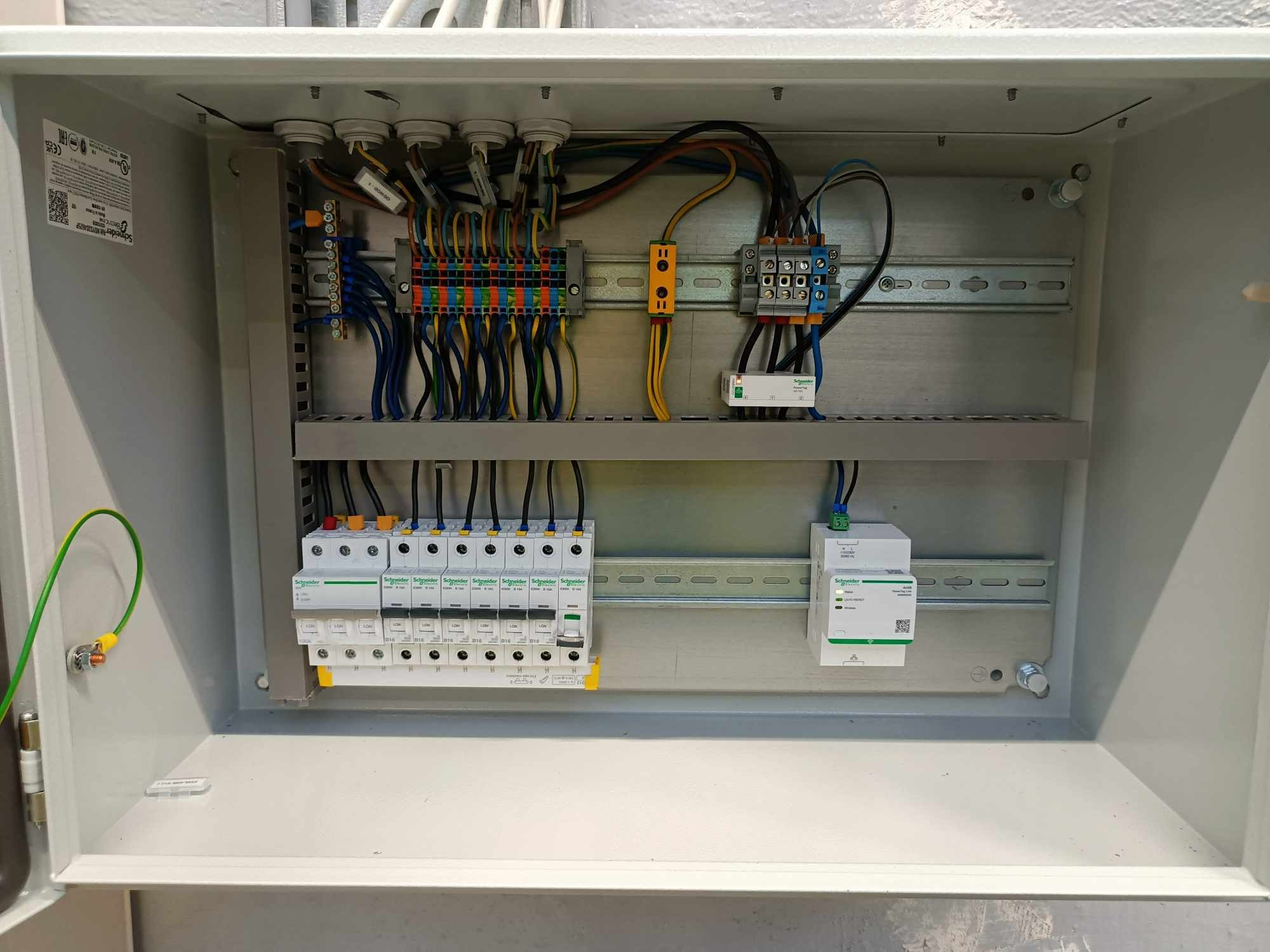 Otwarta skrzynka elektryczna z zainstalowanymi bezpiecznikami, przewodami i elementami zabezpieczającymi. Widoczne złącza, listwy zaciskowe i przewody w różnych kolorach.