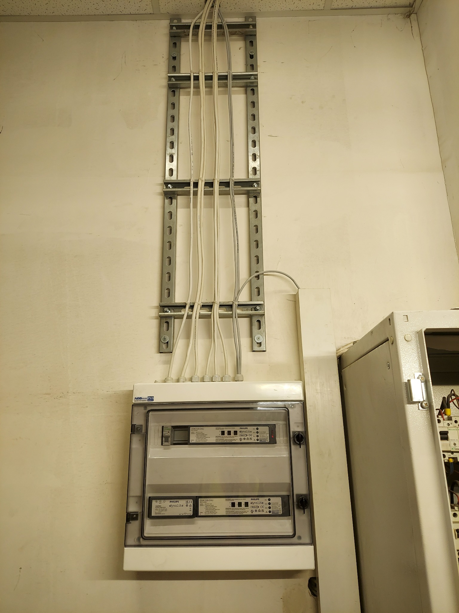 Rozdzielnica elektryczna Philips Dynalite z przewodami w metalowej szynie montażowej na kremowej ścianie, widok z dołu, obok szafa sterownicza.