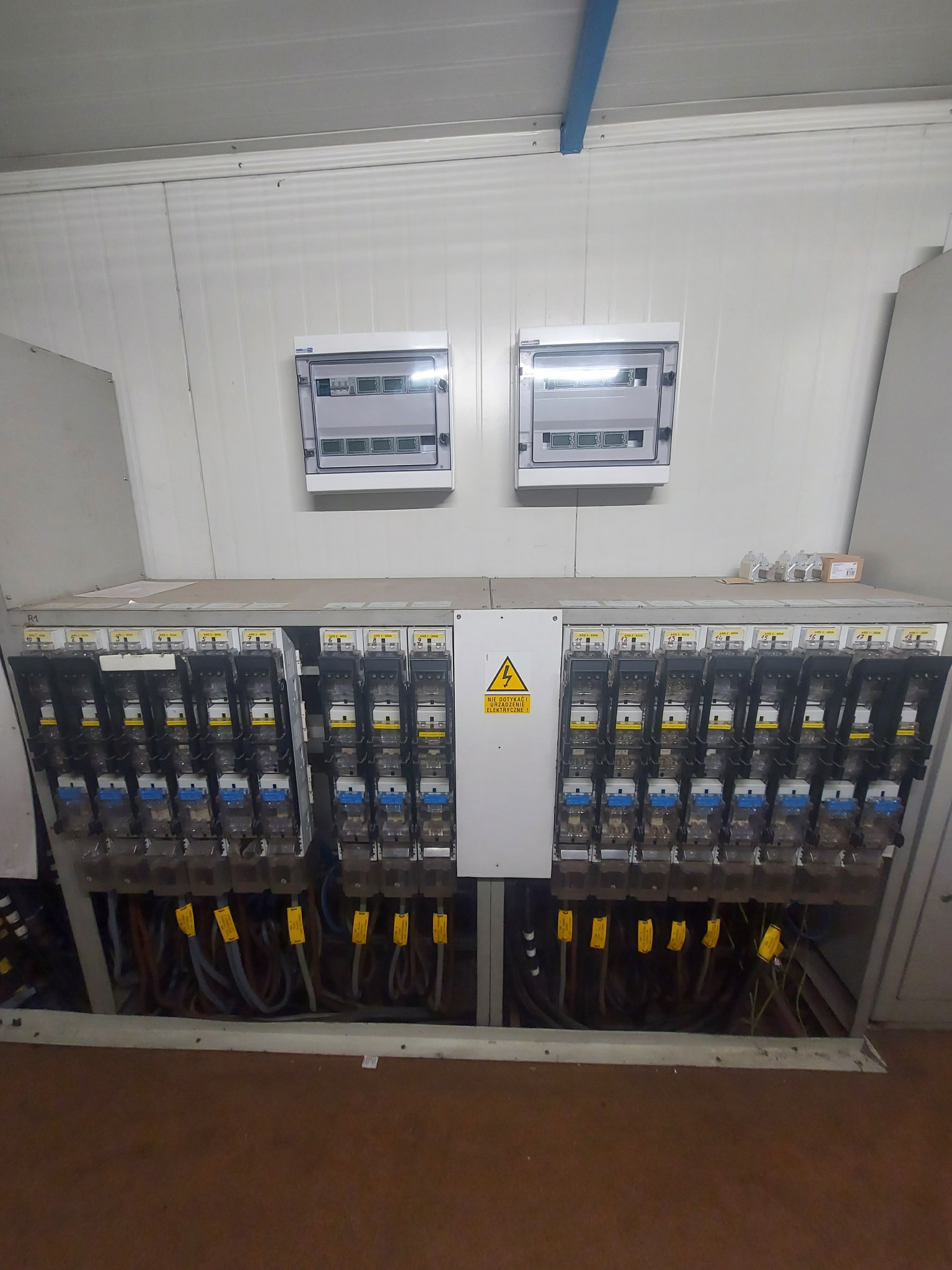 Rozbudowana rozdzielnica elektryczna z licznymi zabezpieczeniami i okablowaniem, uzupełniona o dwie mniejsze skrzynki na ścianie. Widoczny znak ostrzegawczy.