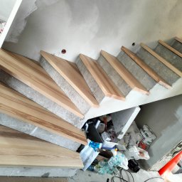 Paweł łuc - Montaż drewnianych stopni schodów na betonowej konstrukcji w surowym wnętrzu. Widoczne materiały budowlane i narzędzia.