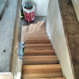 Paweł łuc - Montaż jasnych, drewnianych schodów w surowym wnętrzu. Widoczne materiały izolacyjne i betonowe elementy konstrukcyjne. Ujęcie z góry.