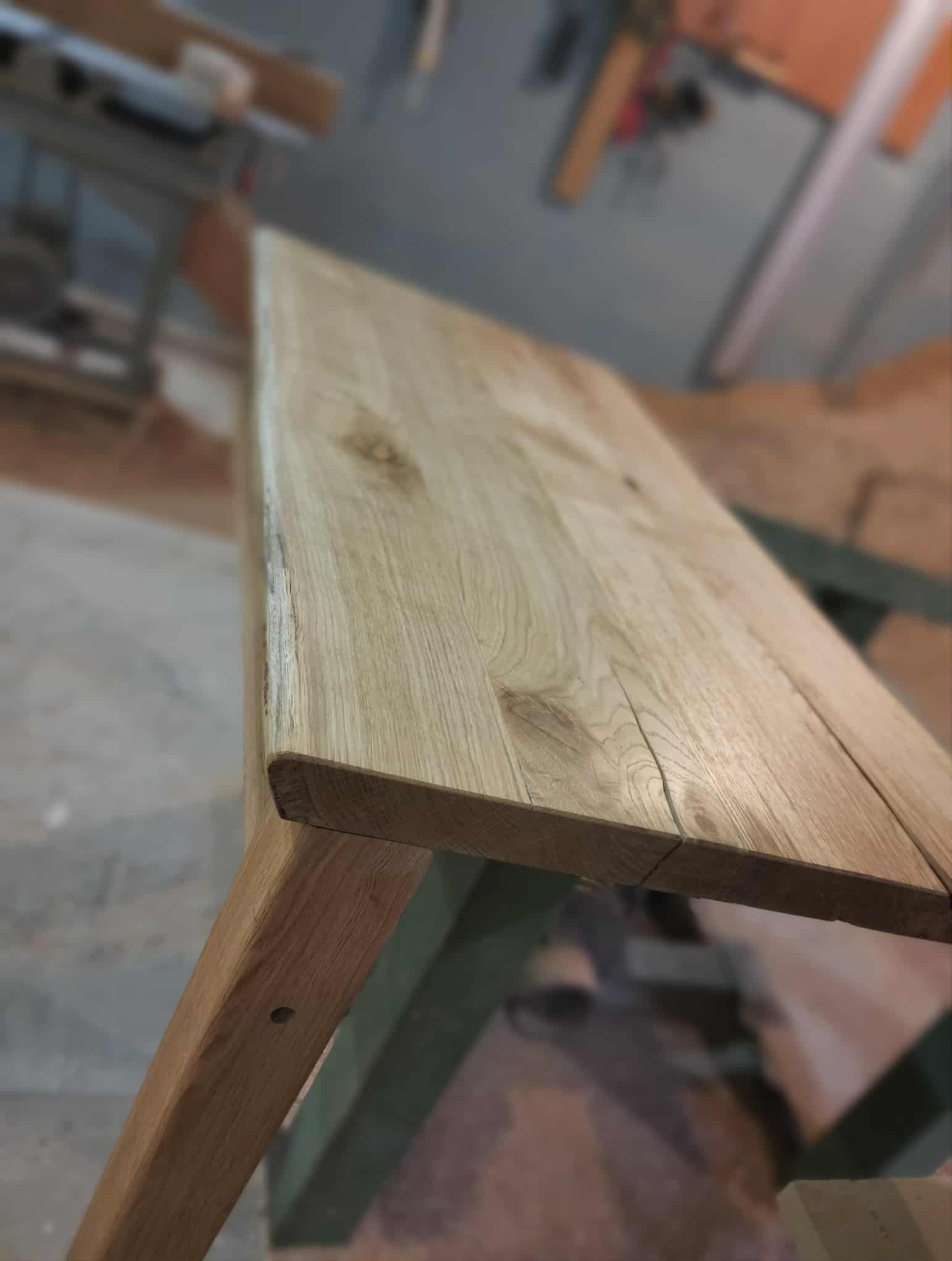 Surowy blat z jasnego drewna dębowego, z widocznymi słojami i naturalnymi pęknięciami, na tle warsztatu stolarskiego. Ujęcie z bliska, ukazujące detale wykończenia.