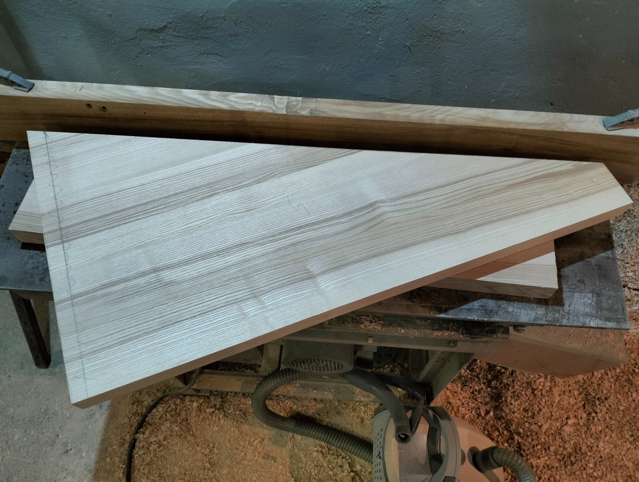 Obróbka drewna: Dwa jasne, trapezoidalne elementy z wyraźnym usłojeniem leżą na metalowym stole warsztatowym, widoczne ślady ołówka i mocowania. W tle szara ściana.