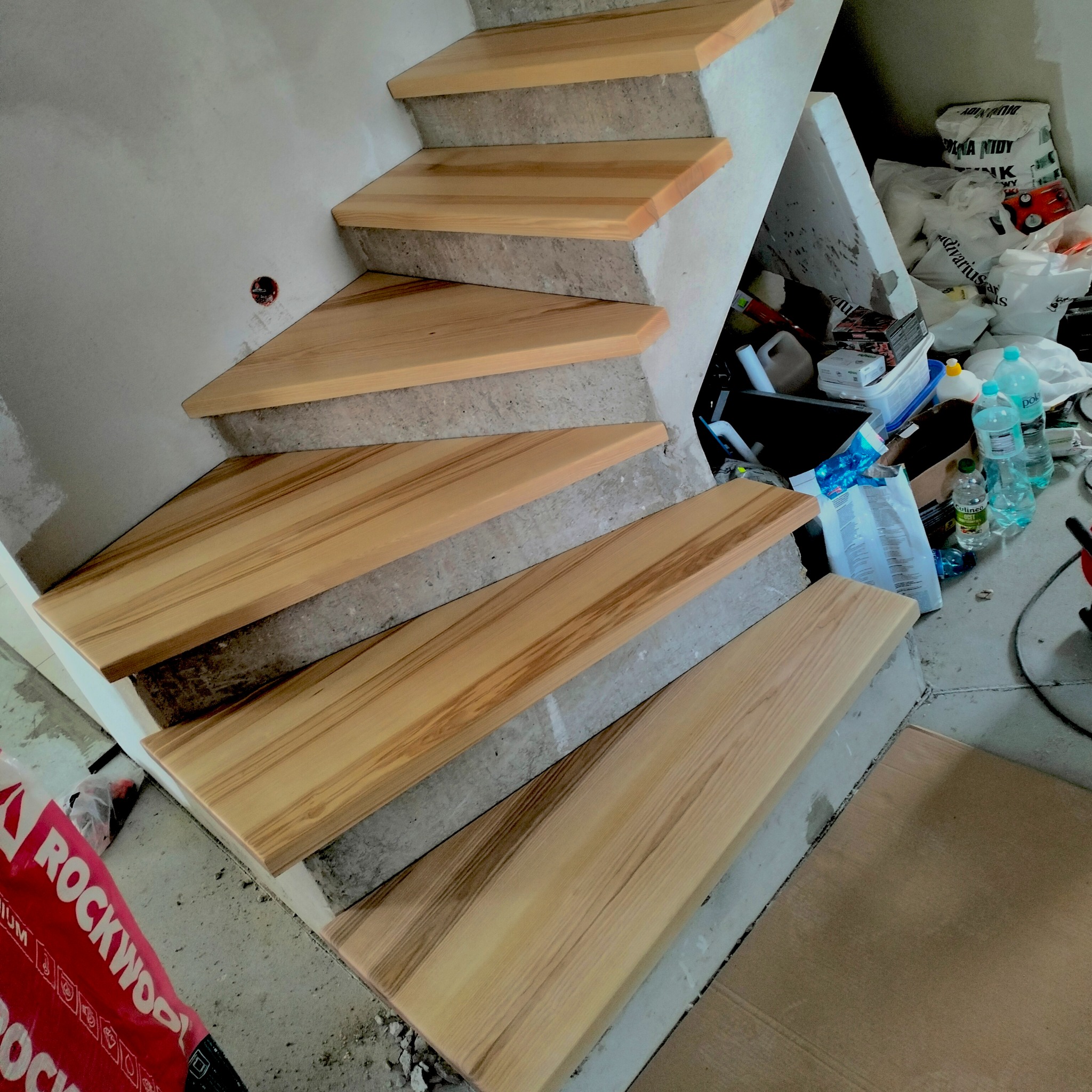 Montaż drewnianych stopni na betonowych schodach w surowym wnętrzu. Widoczne materiały budowlane i narzędzia, co sugeruje etap wykończeniowy.