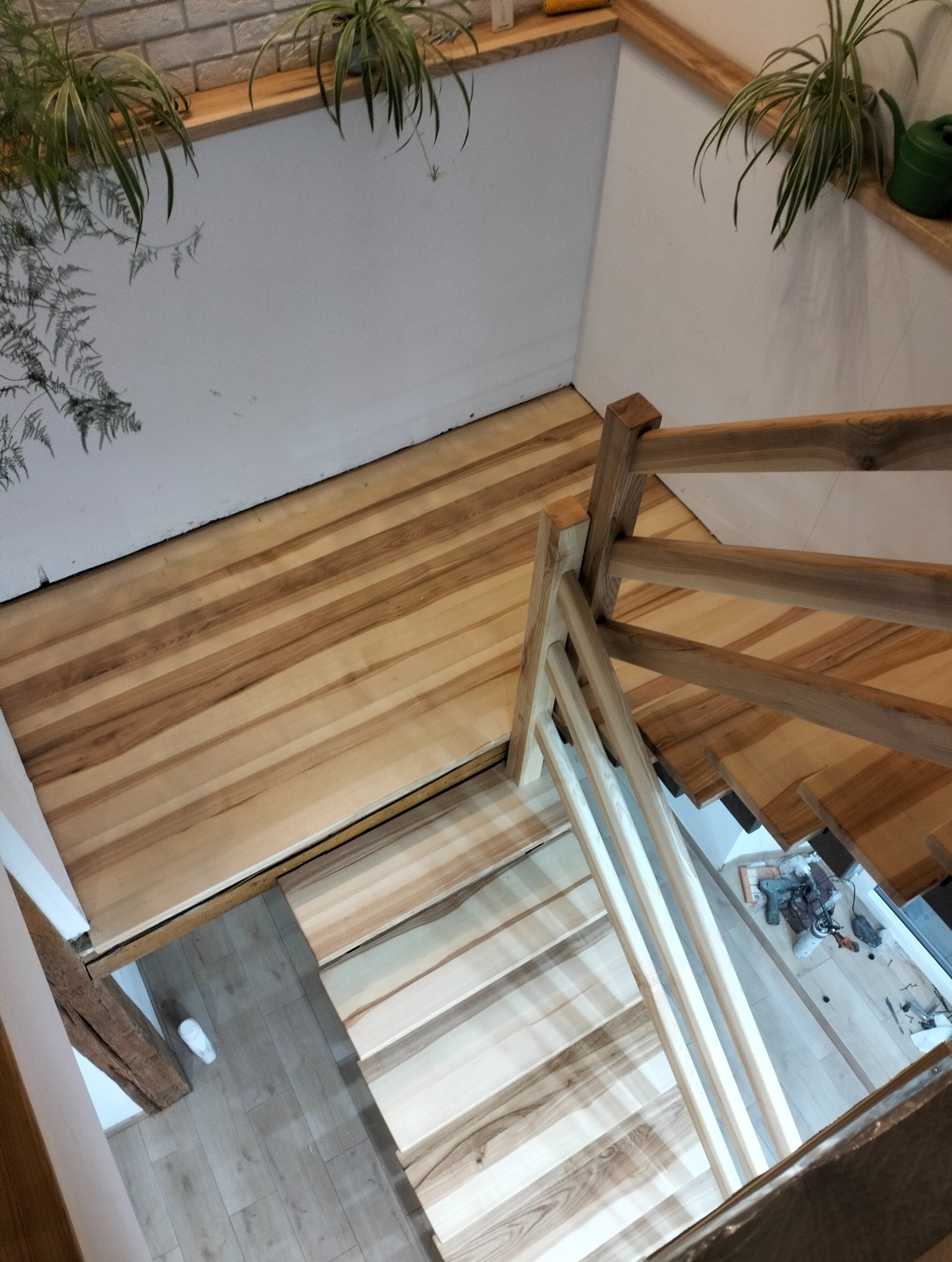 Jasne, drewniane schody z balustradą, widziane z góry. Na ścianach rośliny doniczkowe. Widoczne narzędzia na podeście. Nietypowy kąt ujęcia podkreśla naturalne drewno.