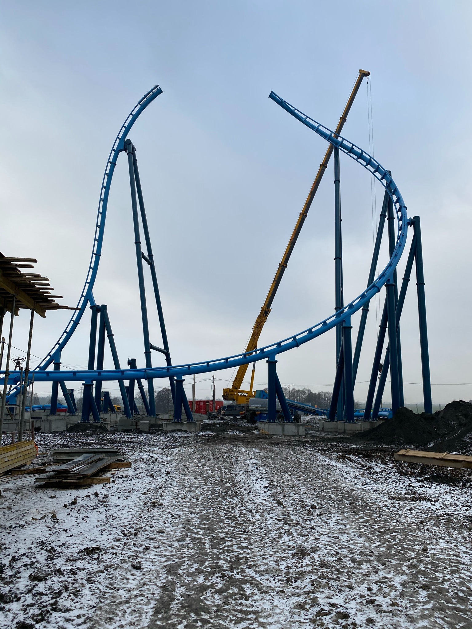 Montaż niebieskiej konstrukcji stalowej, prawdopodobnie kolejki górskiej, z użyciem żółtego dźwigu na placu budowy w pochmurny dzień. Widoczne fundamenty i elementy konstrukcyjne.
