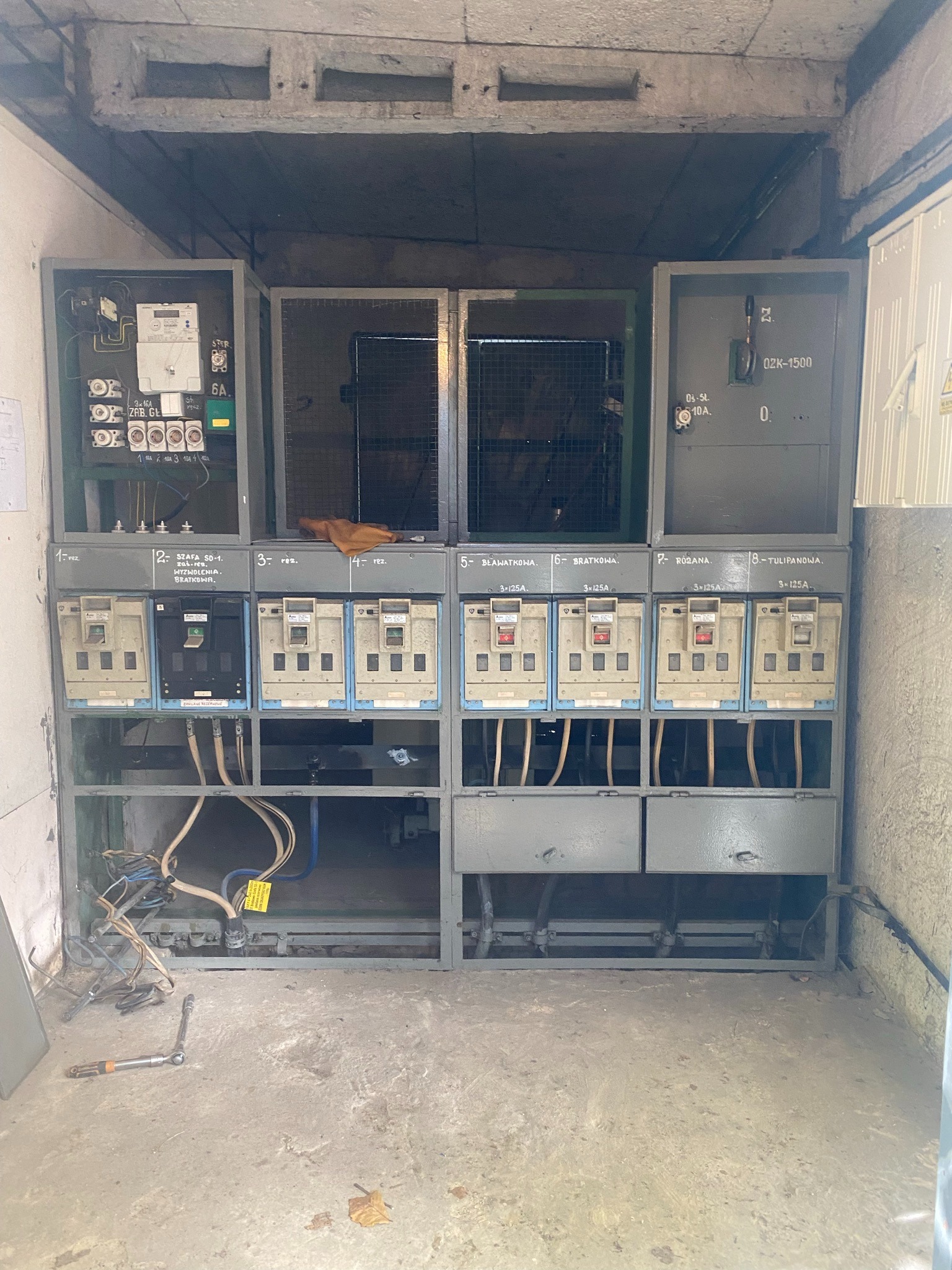 Rozdzielnia elektryczna z bezpiecznikami i licznikami w szarej obudowie, widoczne kable i narzędzia na podłodze. Oświetlenie sufitowe.