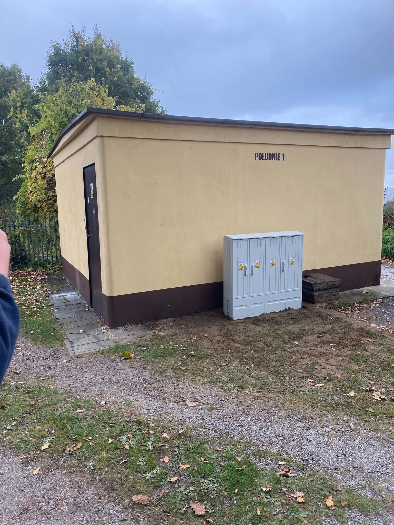 Zewnętrzna stacja transformatorowa z napisem 'Południe 1' i szarą szafą elektryczną na trawiastym terenie. Widoczny fragment dłoni w lewym dolnym rogu.