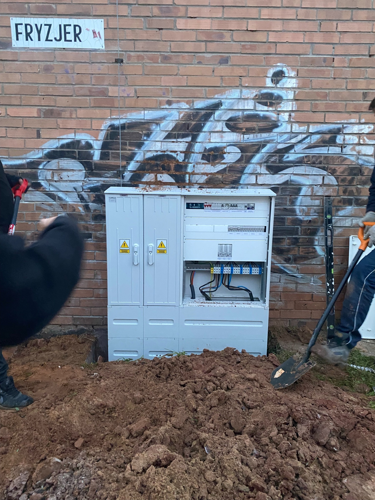 Montaż skrzynki elektrycznej na zewnątrz budynku z ceglaną ścianą i graffiti. Widoczne kable, poziomica i łopata. Tabliczka z napisem 'Fryzjer' w tle.