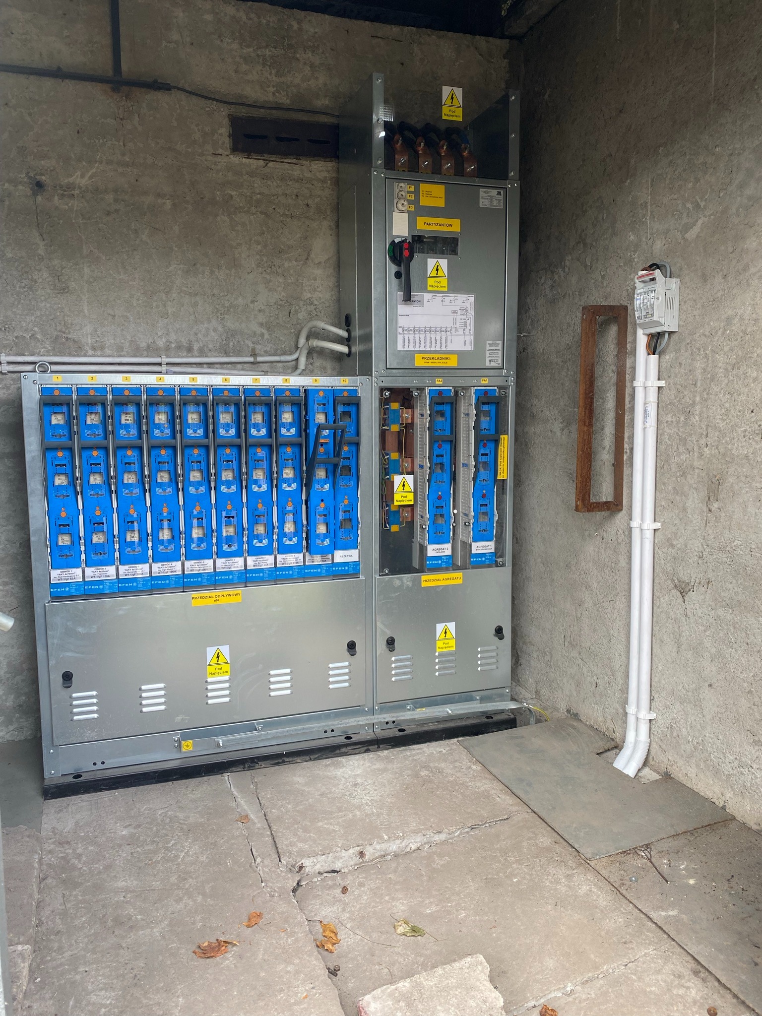 Rozdzielnica elektryczna z bezpiecznikami i oznaczeniami ostrzegawczymi, zamontowana w surowym, betonowym pomieszczeniu. Widoczne przewody i elementy instalacji.