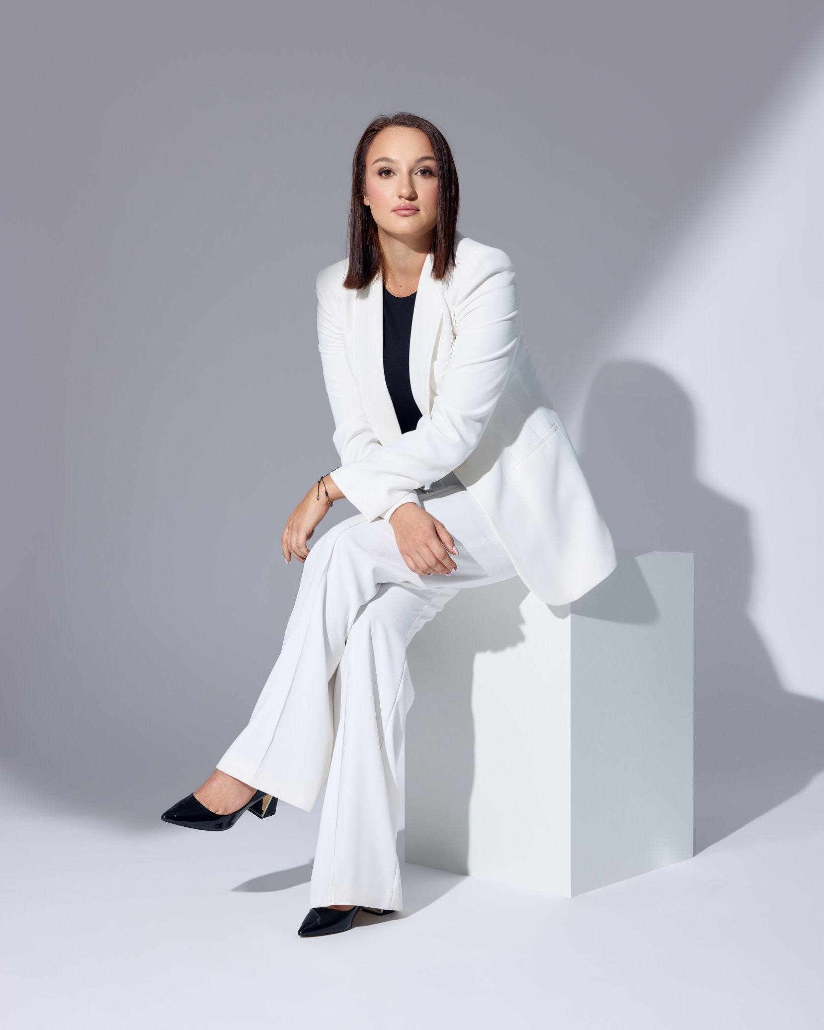 Kobieta w białym garniturze siedzi na białym sześcianie, patrząc w obiektyw. Profesjonalny wizerunek w minimalistycznym studio z subtelnym oświetleniem.