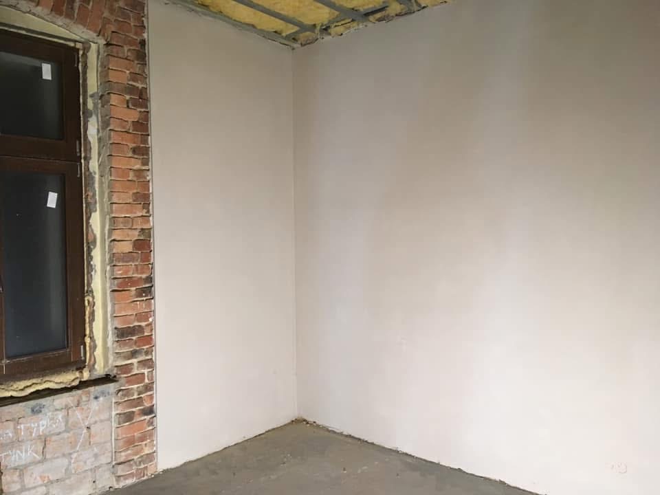 Gładka, jasna ściana po nałożeniu tynku maszynowego, widoczna obok okna z ceglaną obudową w surowym stanie. Sufit z widoczną izolacją.