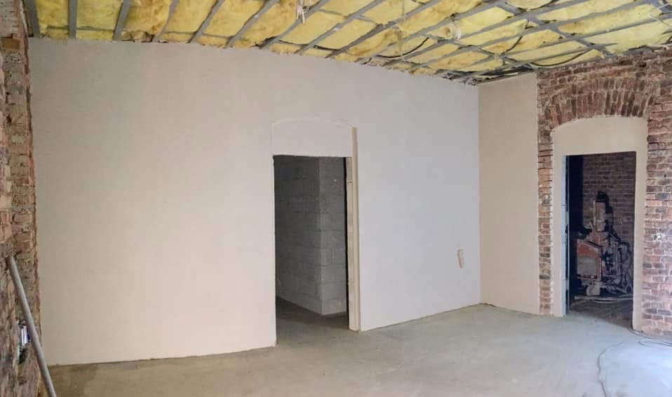 Wnętrze po tynkowaniu maszynowym: gładkie, jasne ściany kontrastują z fragmentami cegły. Widoczne ocieplenie sufitu i otwory drzwiowe. Podłoga betonowa, surowy stan.